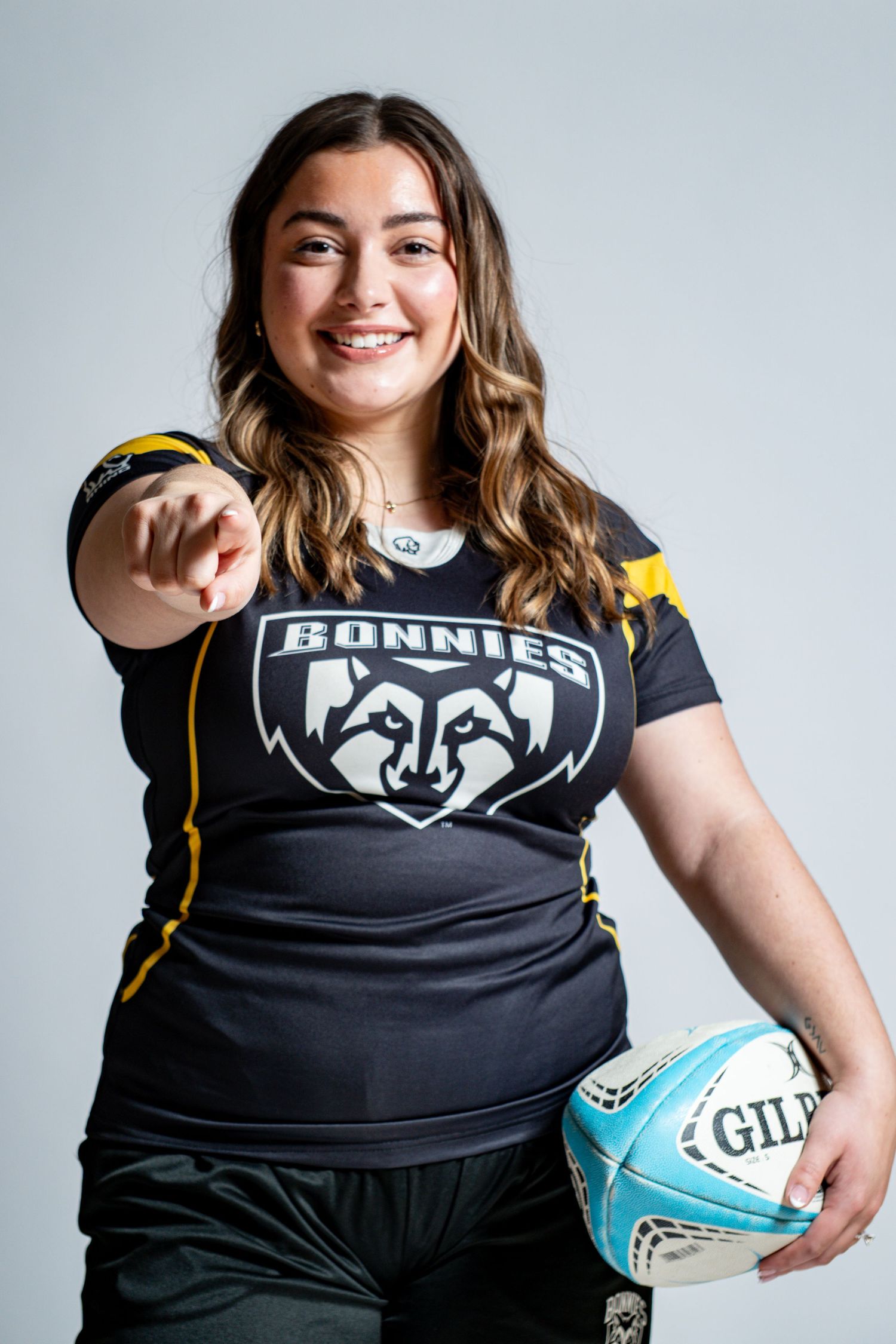 Smiling rugby player in a black Bonnies jersey points toward the camera while holding a light blue Gilbert rugby ball at her side.