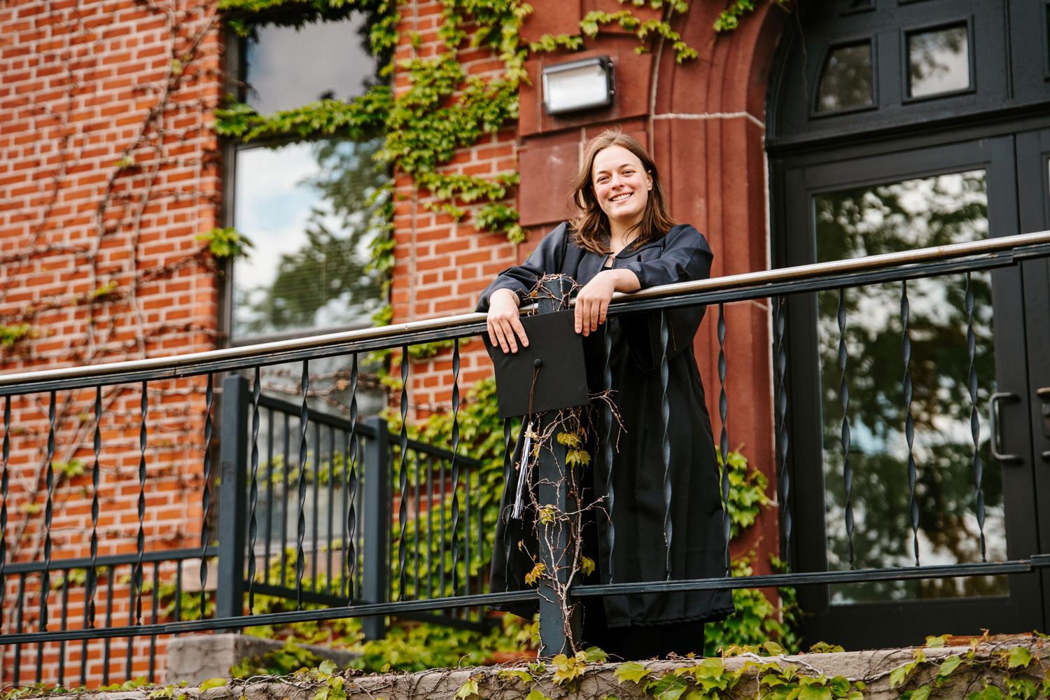 Smiling graduate in a black gown leans on a railing outside an ivy-covered brick building, holding her cap.