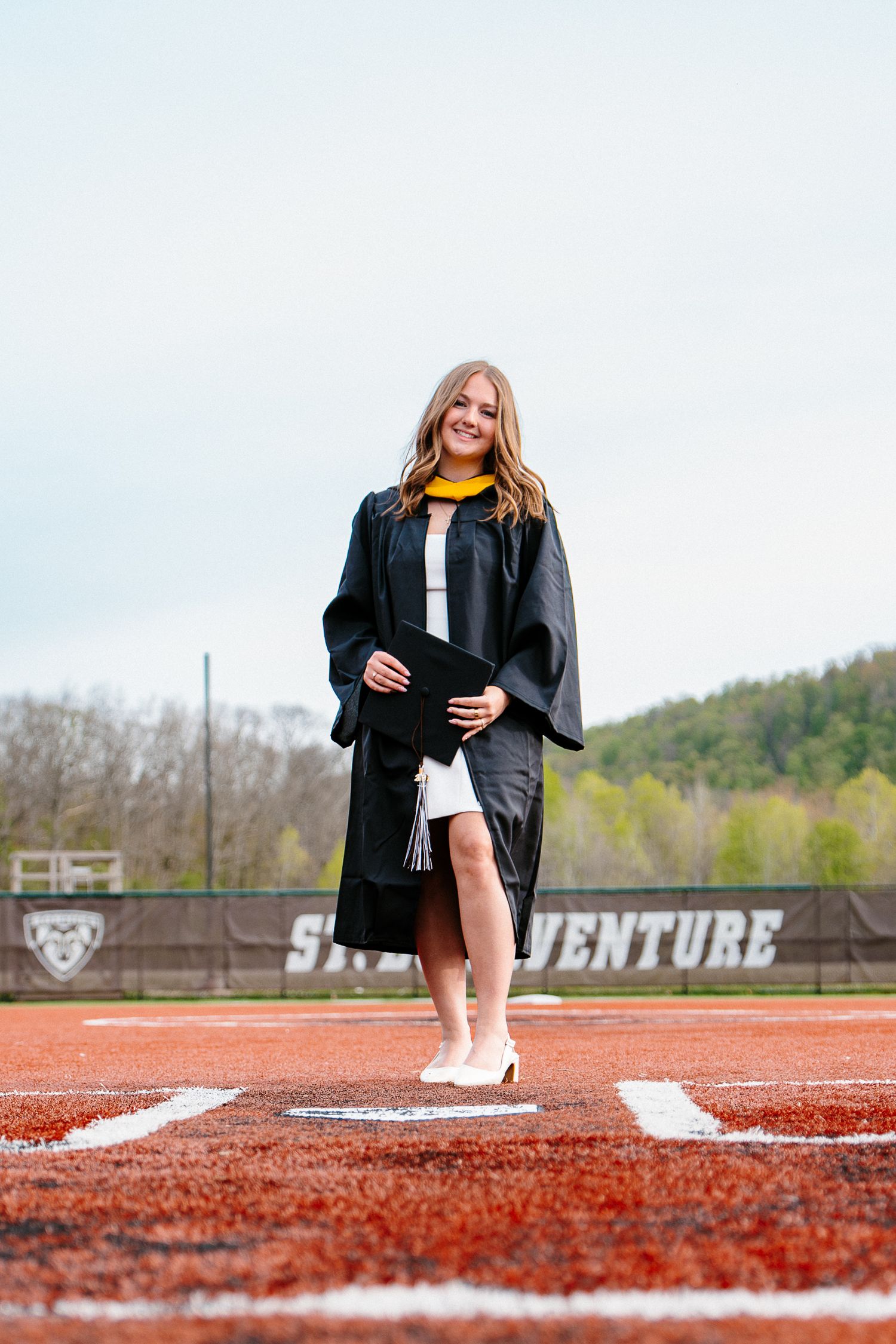 Graduate in a black gown with a gold hood holds her cap while standing on a softball field with green hills behind her.