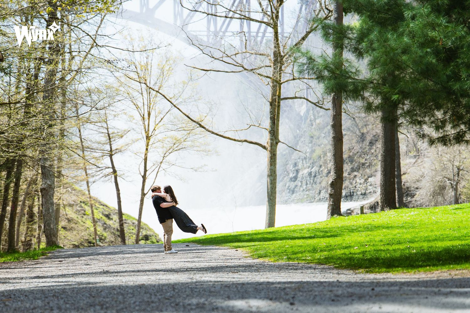 A couple embraces and twirls on a gravel path beside a green lawn and tall trees in a scenic spring engagement photo.
