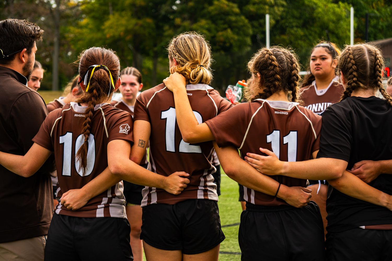 Women's rugby team in brown jerseys huddles with arms linked on a grass field while a coach stands beside them.