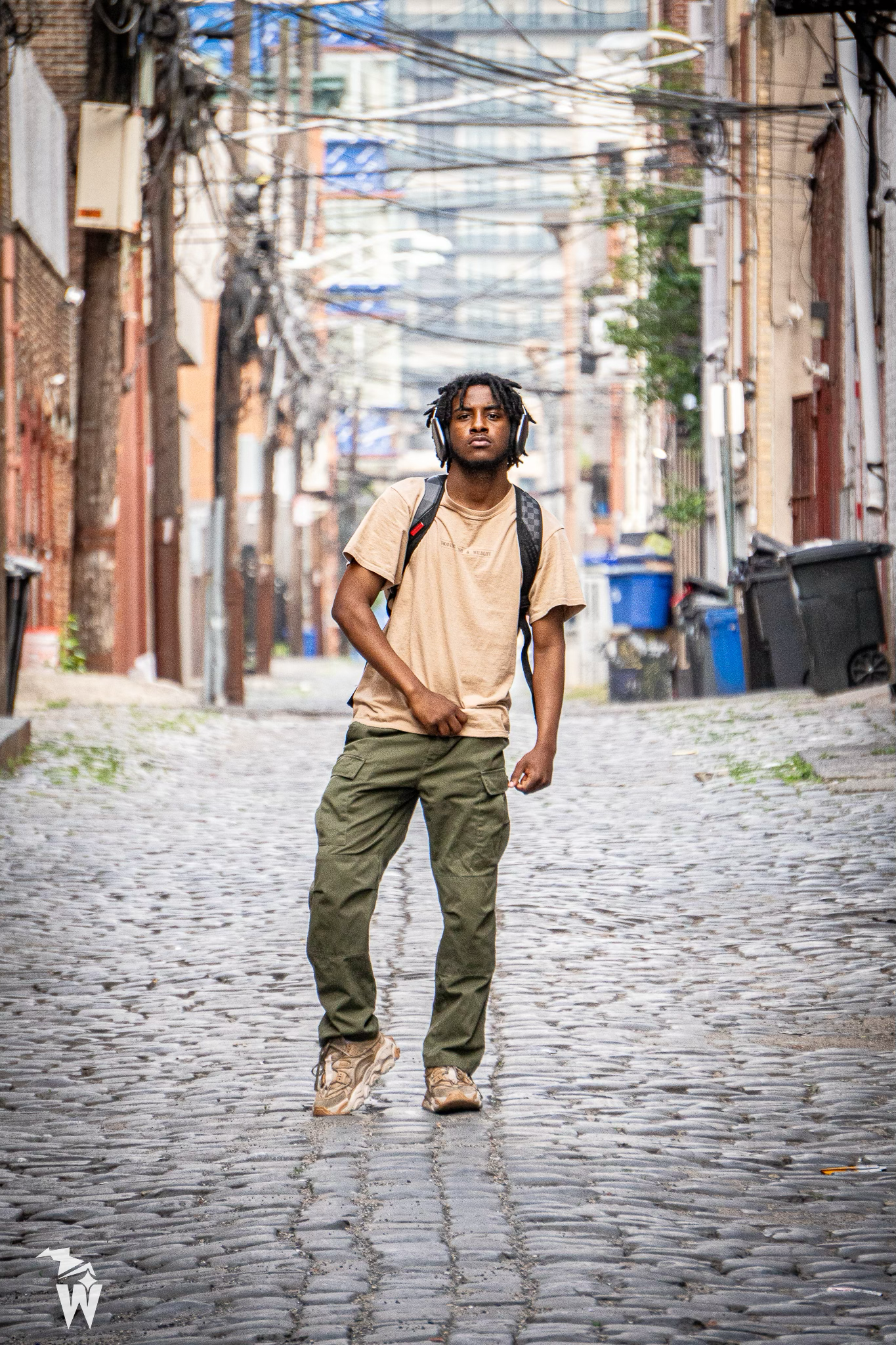 Young man with headphones and a backpack walks down a cobblestone alley lined with brick buildings and utility wires.
