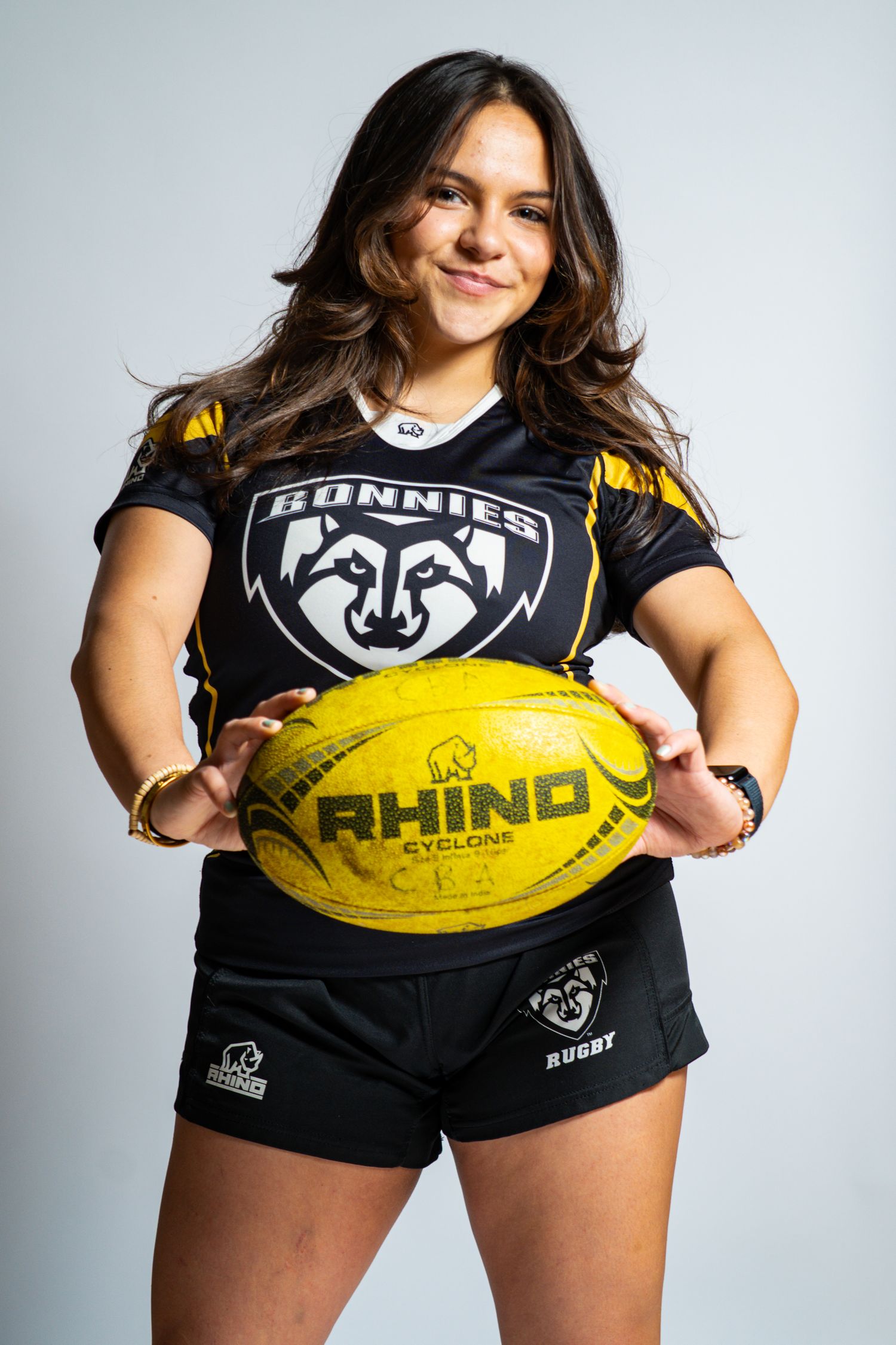 Rugby player in a black Bonnies jersey and shorts holds a yellow Rhino Cyclone rugby ball at her waist with a slight smile.