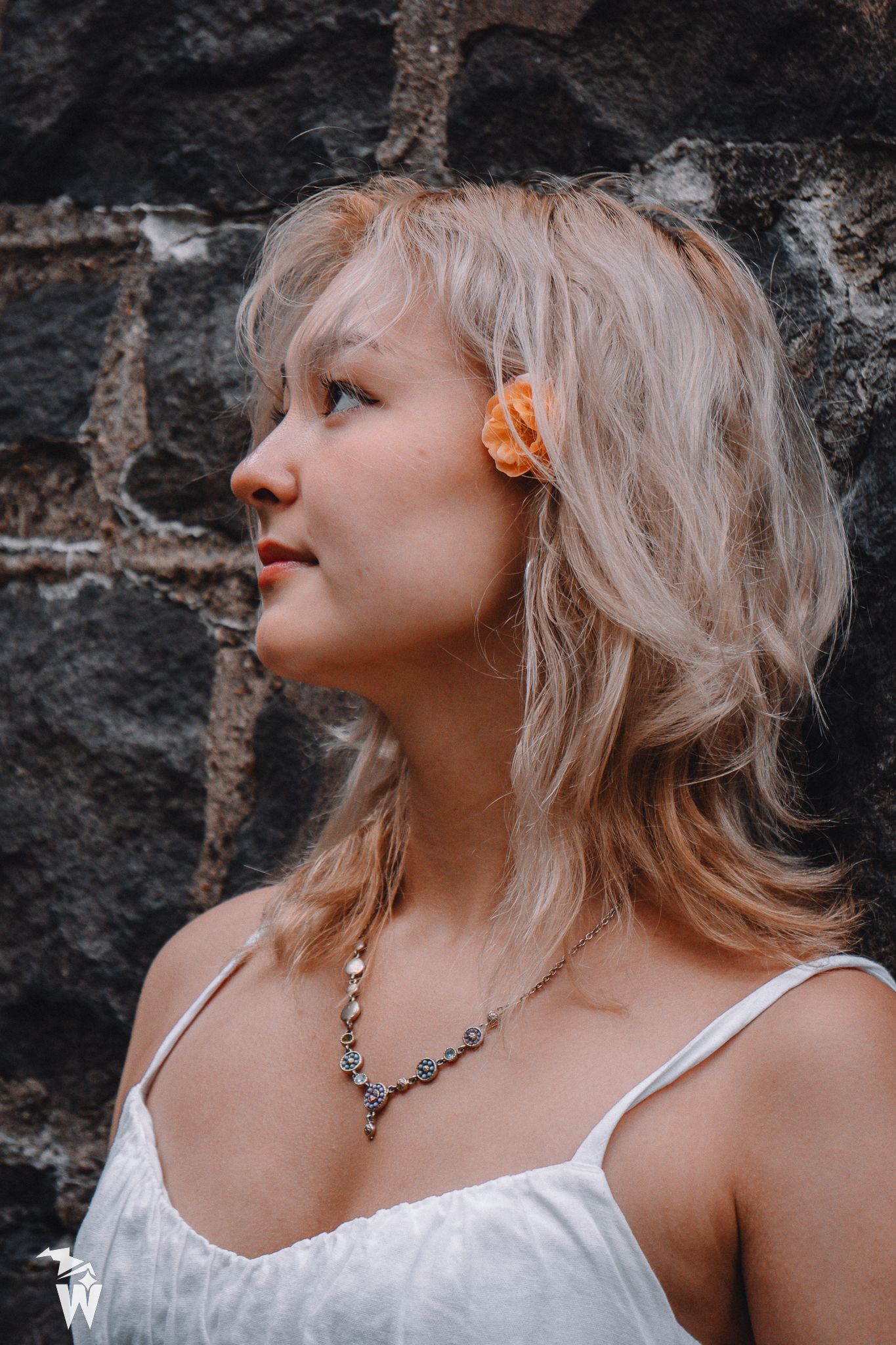 Blonde woman with an orange flower in her hair and a beaded necklace gazes aside against a dark stone wall in a white camisole.