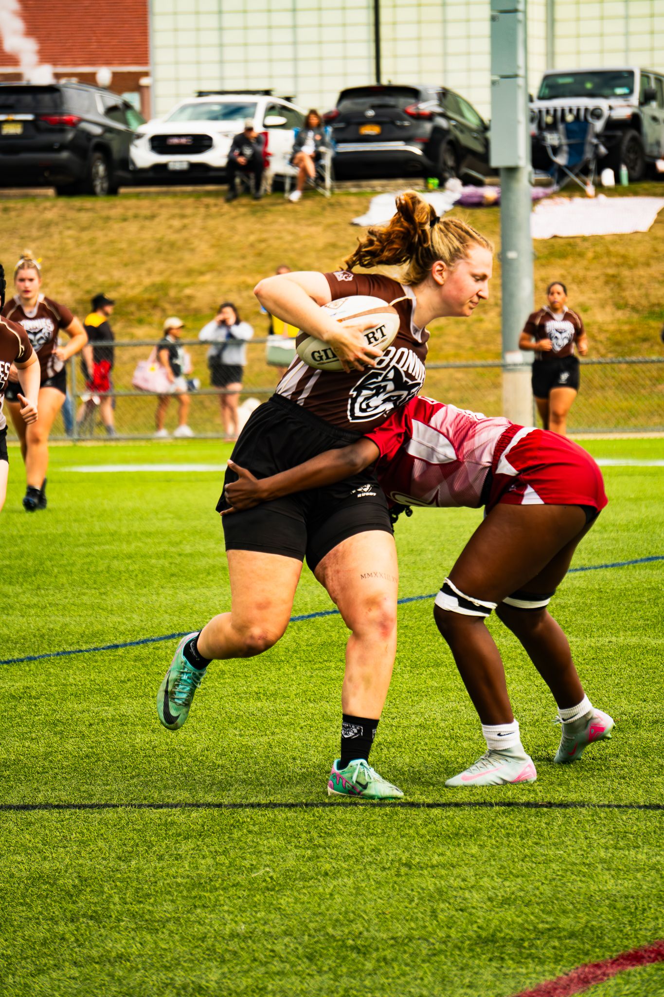 A rugby player in a maroon jersey carries the ball and braces as a defender in a red uniform reaches to tackle her.