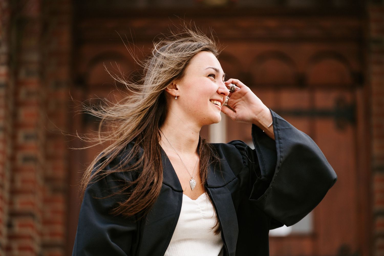 Graduate in a black gown laughs as wind blows her long hair, standing in front of a red brick building.