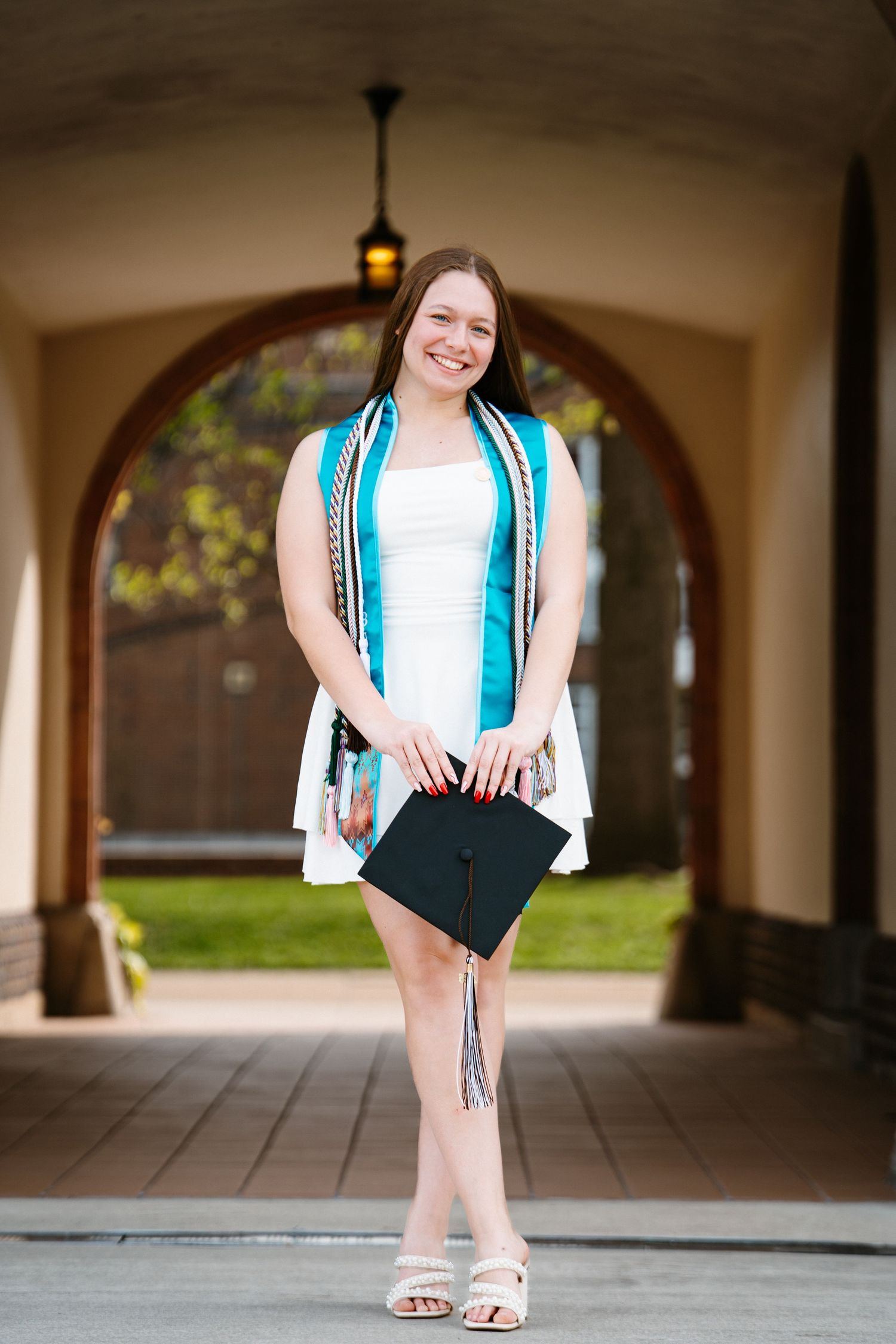 Smiling graduate in a white dress and teal stole holds her black cap with a tassel while standing in a sunlit archway.