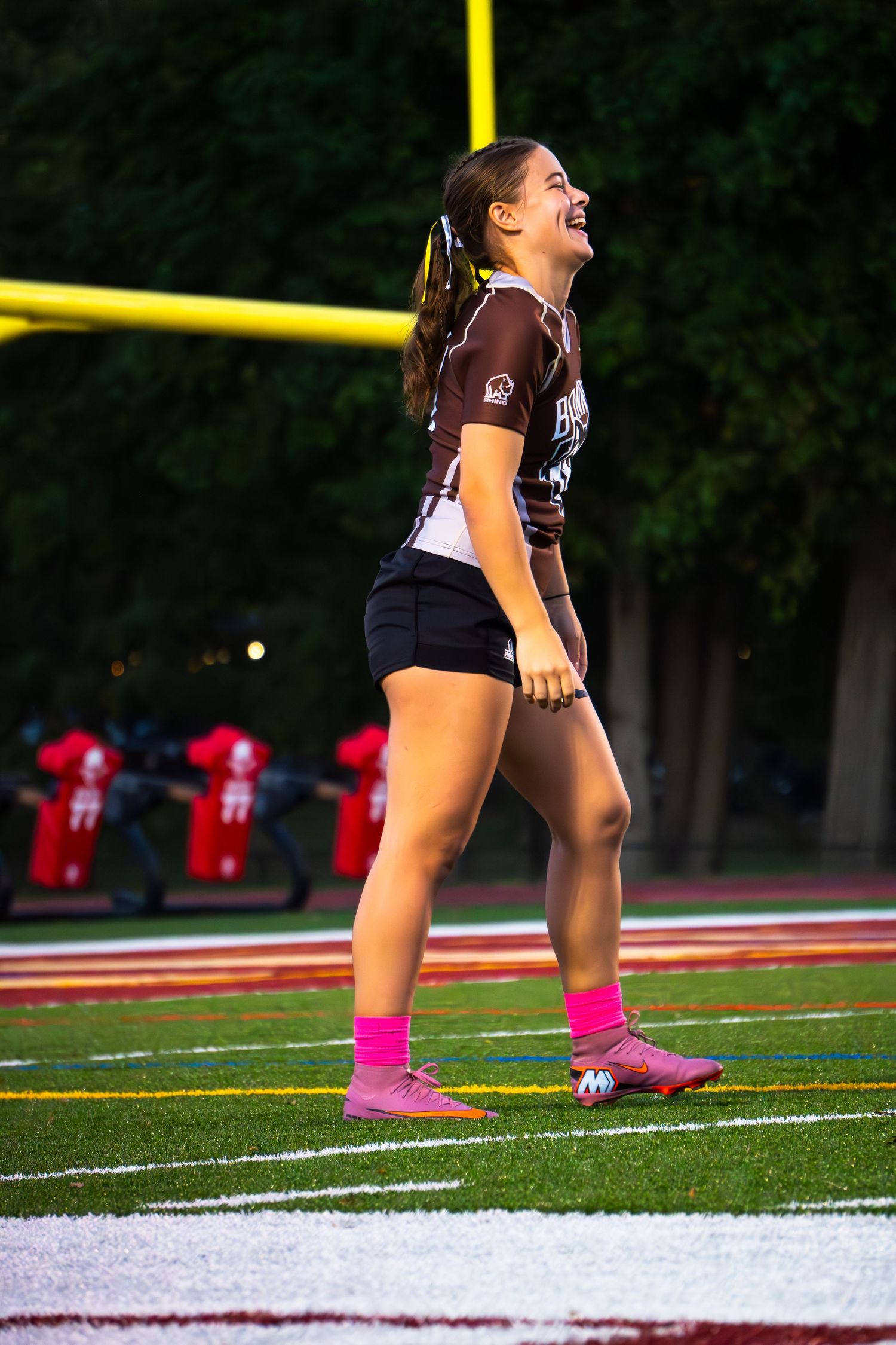Smiling girl in a brown jersey, black shorts and pink socks laughs on a football field near yellow goalposts at dusk.