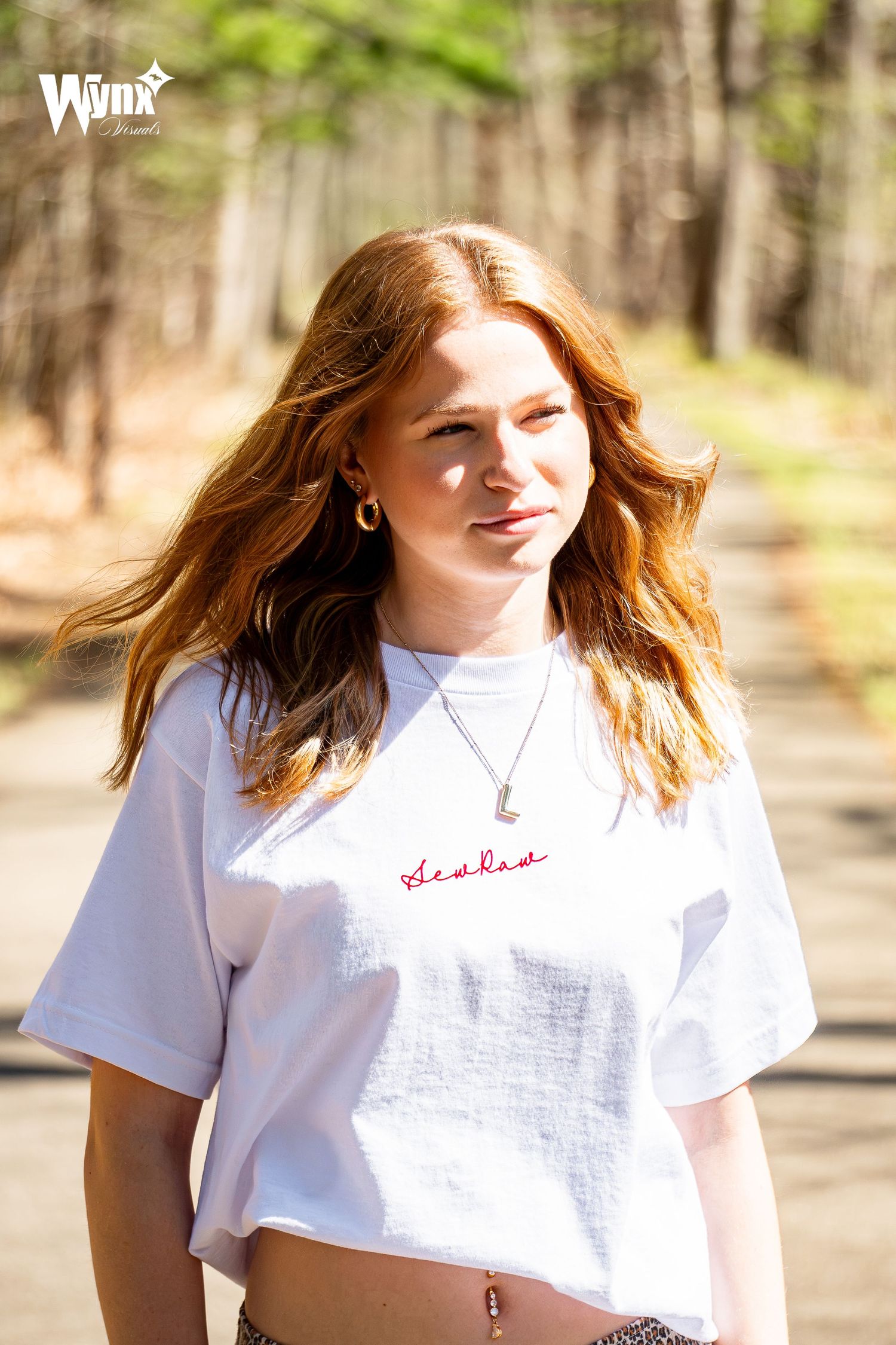 Woman with long red hair wearing a white tee with red Sewlaw script and a pendant necklace, walking along a sunlit path.