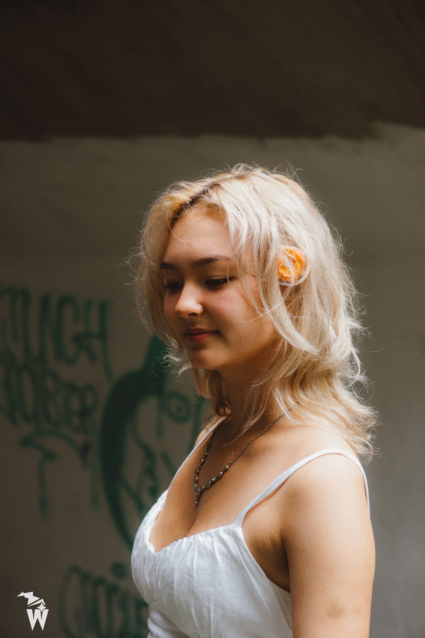 Young woman with blonde hair and an orange flower in a white sundress standing in profile before a graffiti-covered wall.