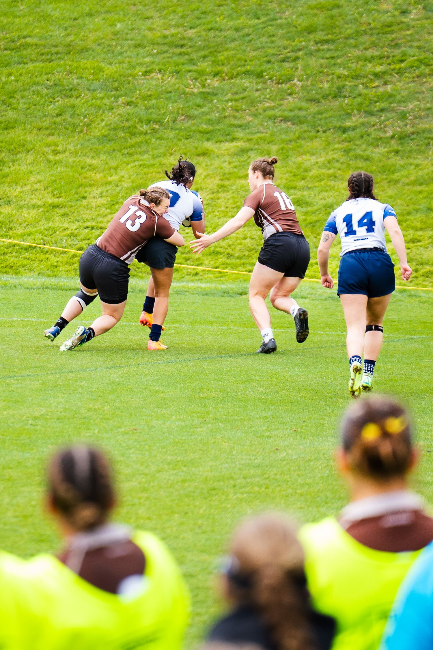 Number 13 in a brown jersey tackles a player in white during a women's rugby match on a green field as teammates look on.