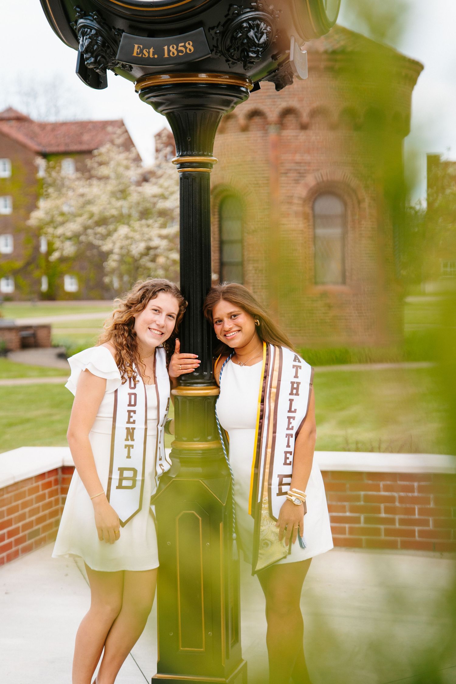 Two smiling graduates in white dresses and Student Athlete stoles pose beside a black Est. 1858 campus clock near brick buildings.