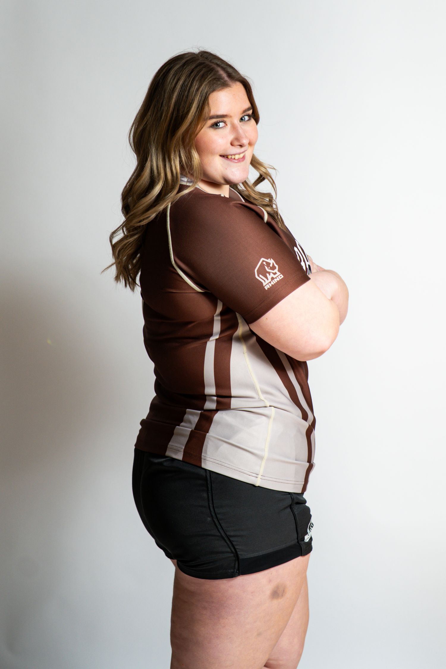 Smiling rugby player with wavy hair poses in profile with folded arms, wearing a brown and tan team jersey and black shorts.