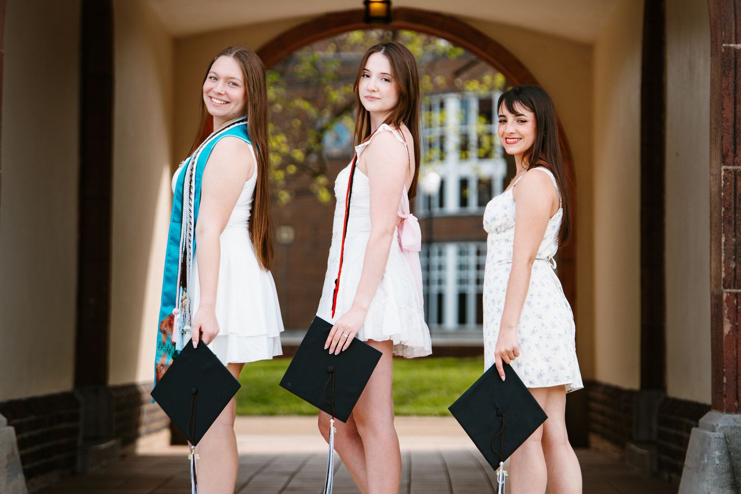 Three smiling graduates in white dresses stand together in a brick archway, each holding a black graduation cap.