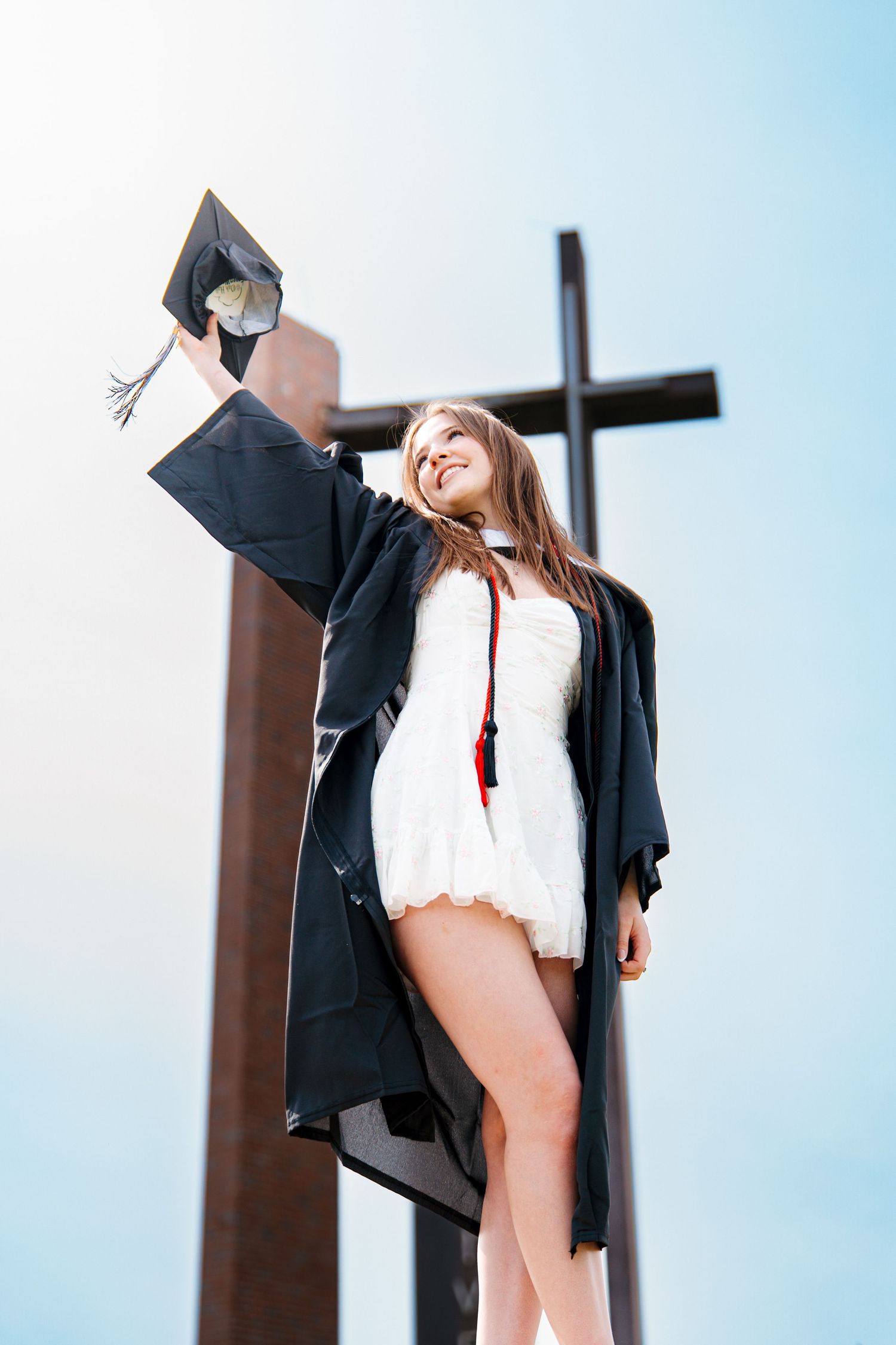 Smiling graduate in a black gown and white dress raises her cap in the air in front of a large cross against a blue sky.
