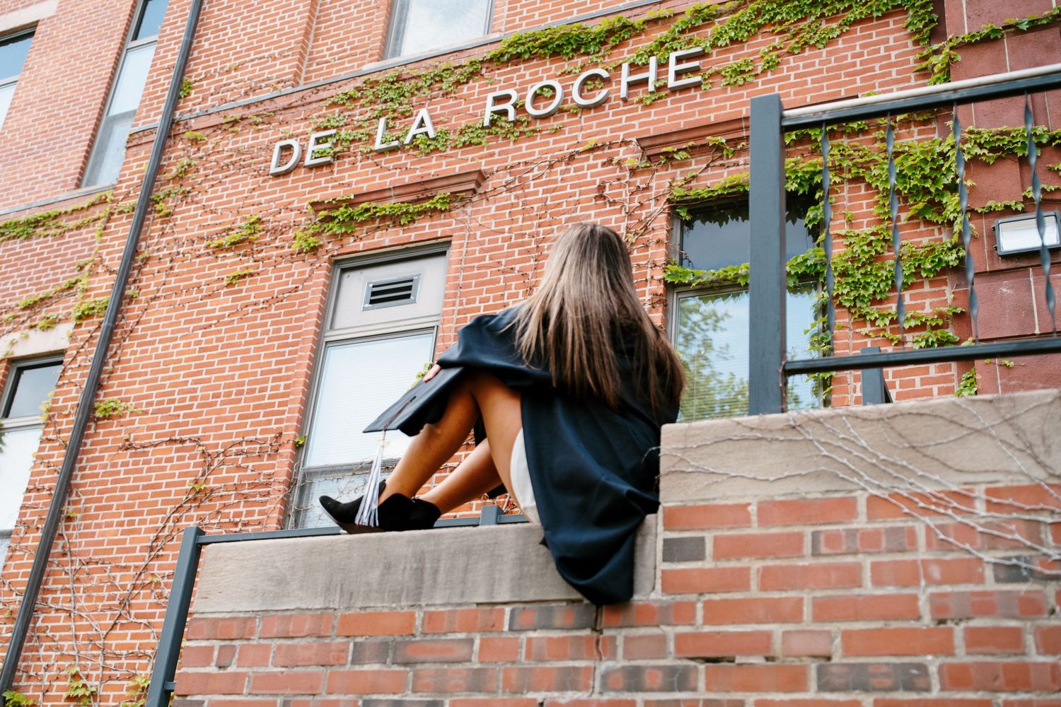 Graduate in a black gown and boots sits on a brick ledge in front of the De La Roche building with ivy-covered walls.