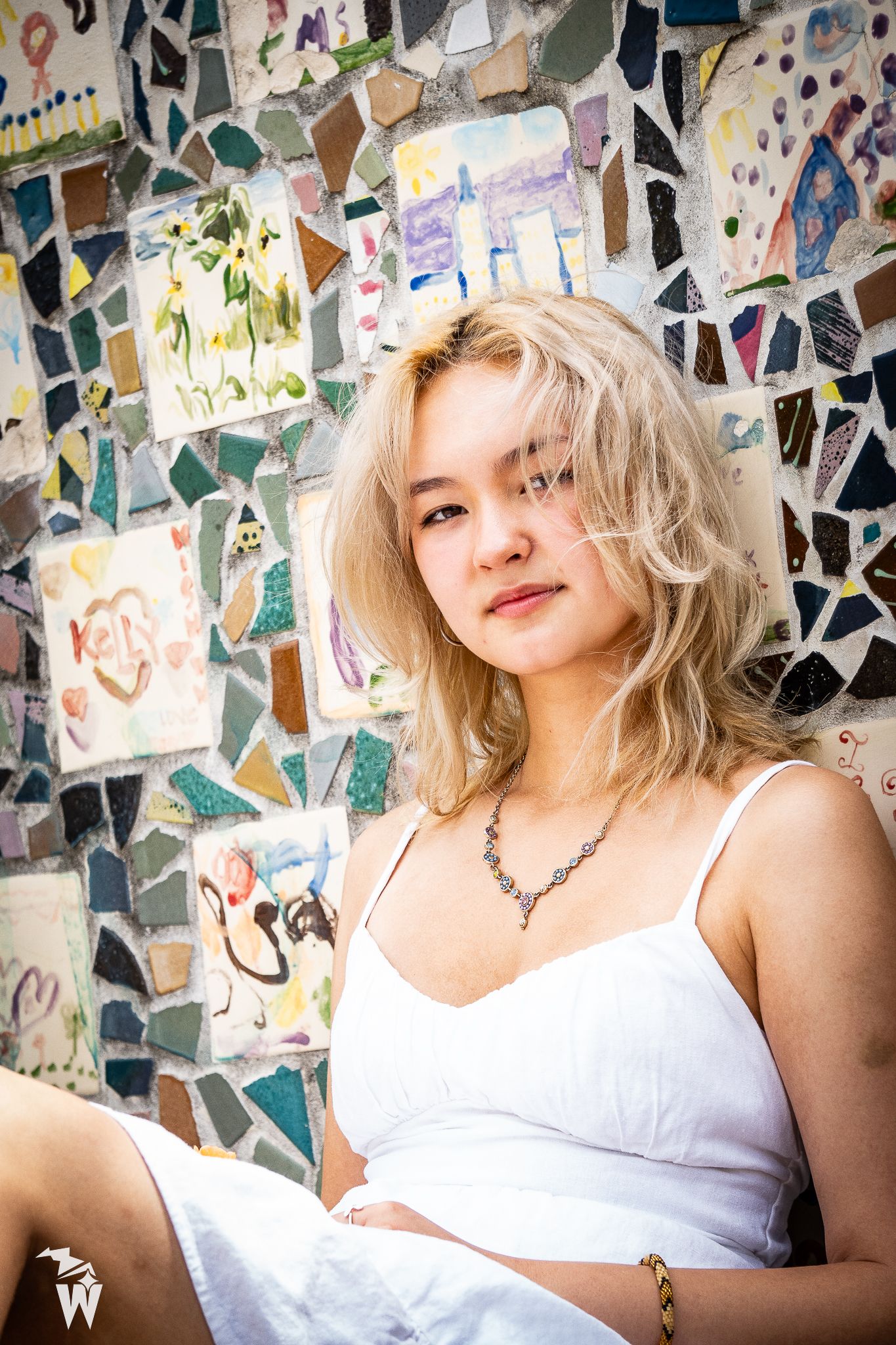 Young woman with tousled blonde hair in a white dress sitting in front of a colorful mosaic tile wall.