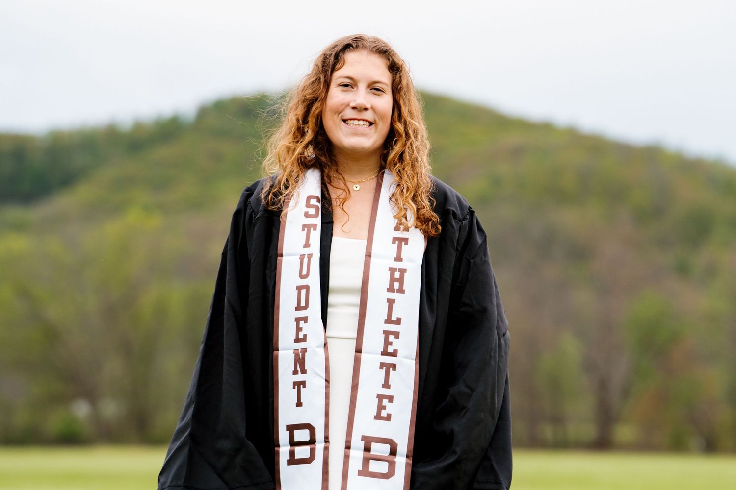 A smiling graduate in a black gown and brown and white Student Athlete stole stands in a grassy field with green hills behind.