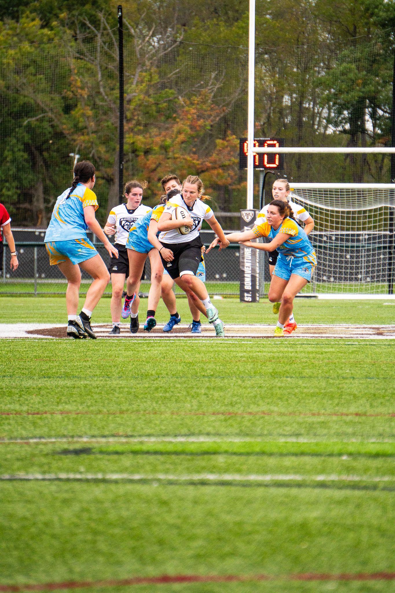 Women's rugby players in white and blue jerseys run and tackle on a turf field near the goalposts on an autumn day.