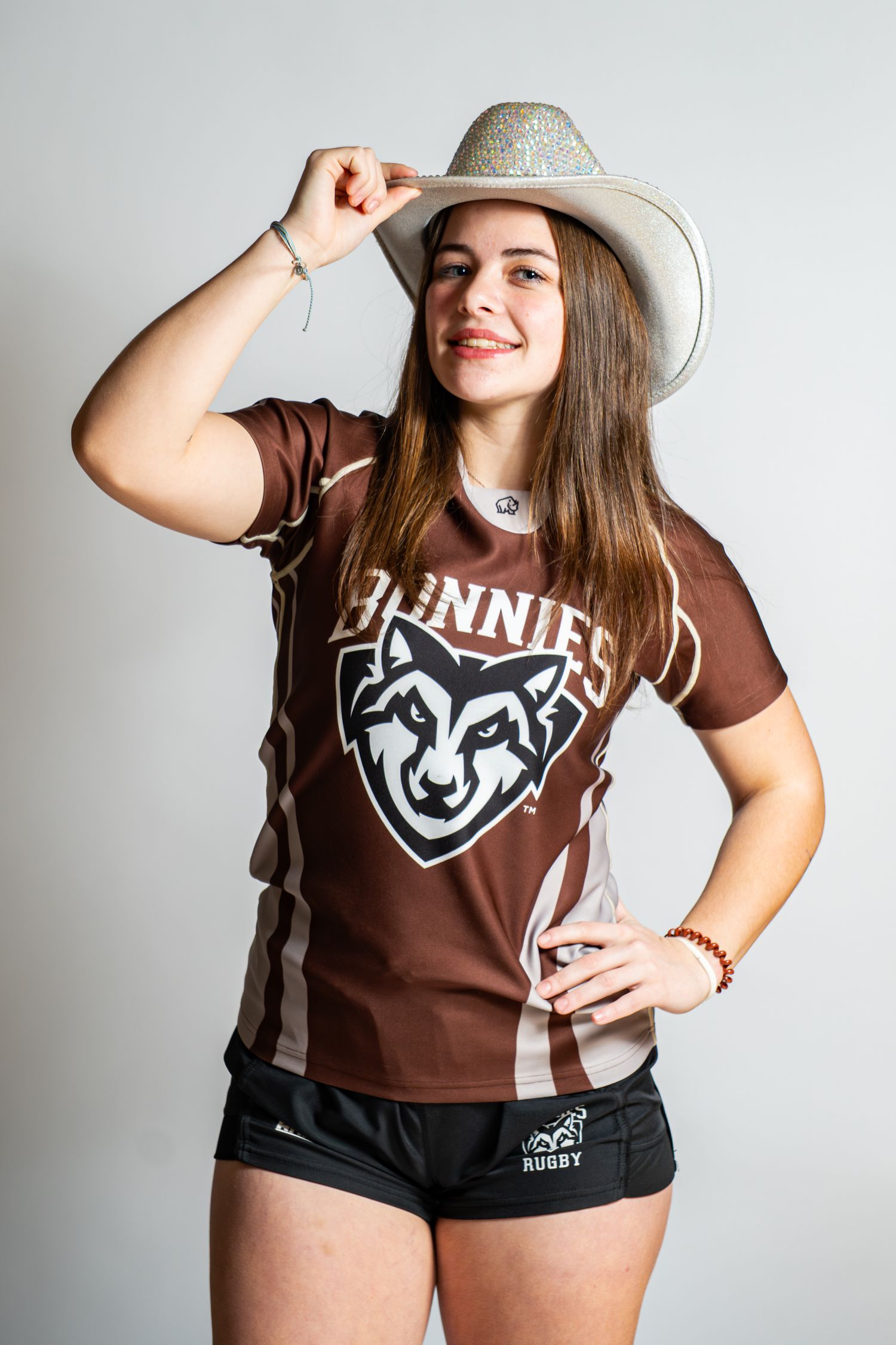 Smiling rugby player in a brown Bonnies jersey and black shorts tips a sparkly white cowboy hat against a plain studio backdrop.