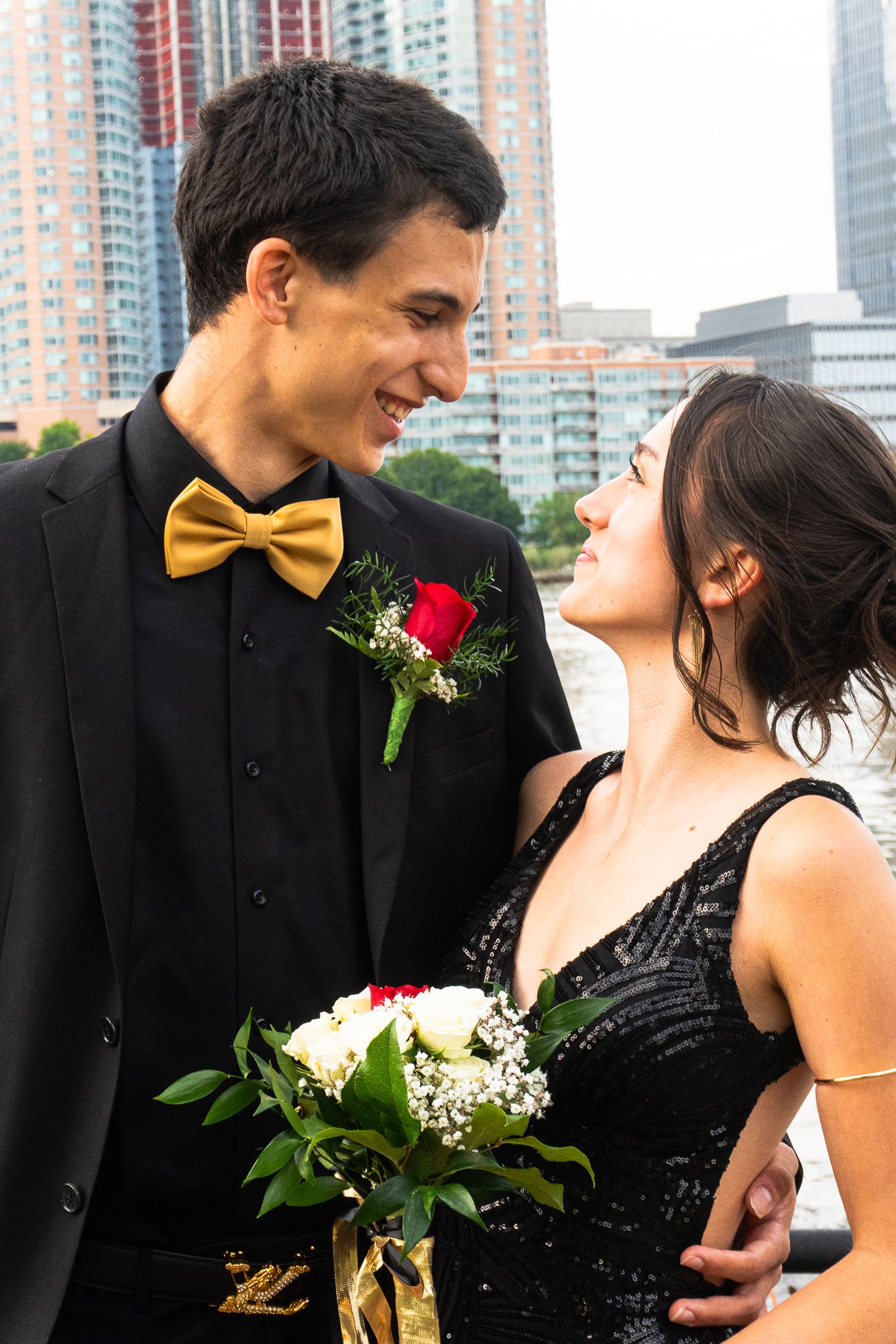 Smiling young man with a gold bow tie and red boutonniere gazes at a woman in a black sequined dress holding white roses.