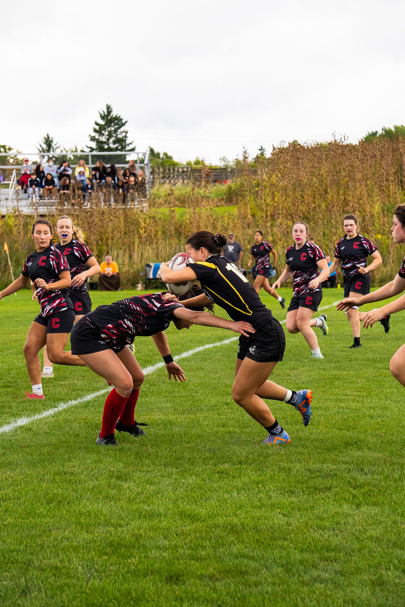A women's rugby ball carrier in a black jersey is tackled by a defender in maroon and black as teammates and spectators look on.