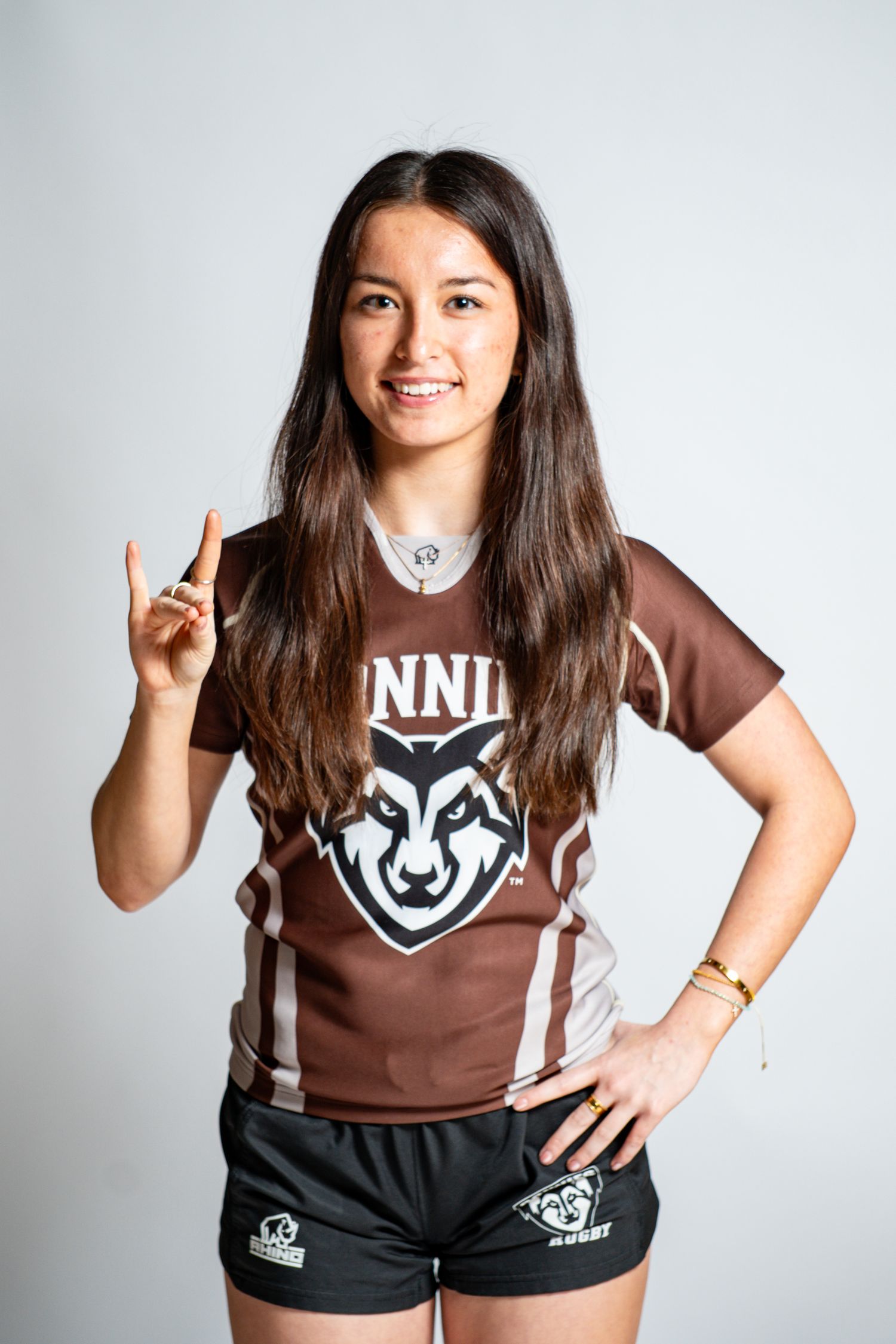 Smiling rugby player with long dark hair in a brown Bonnies jersey makes a hand gesture while resting her other hand on her hip.