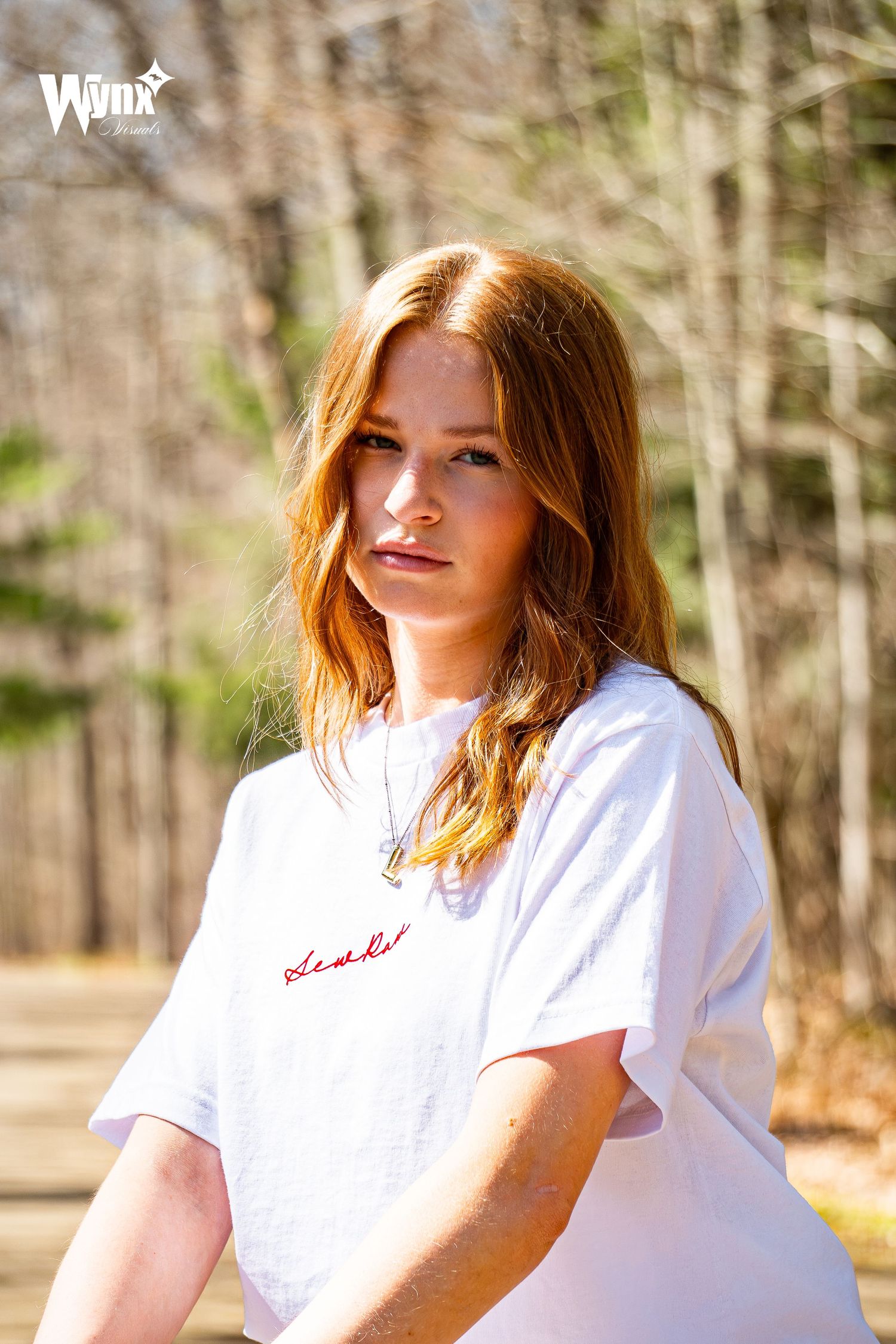 Smiling red-haired woman laughing in a white embroidered t-shirt and pendant necklace on a sunny wooded trail.
