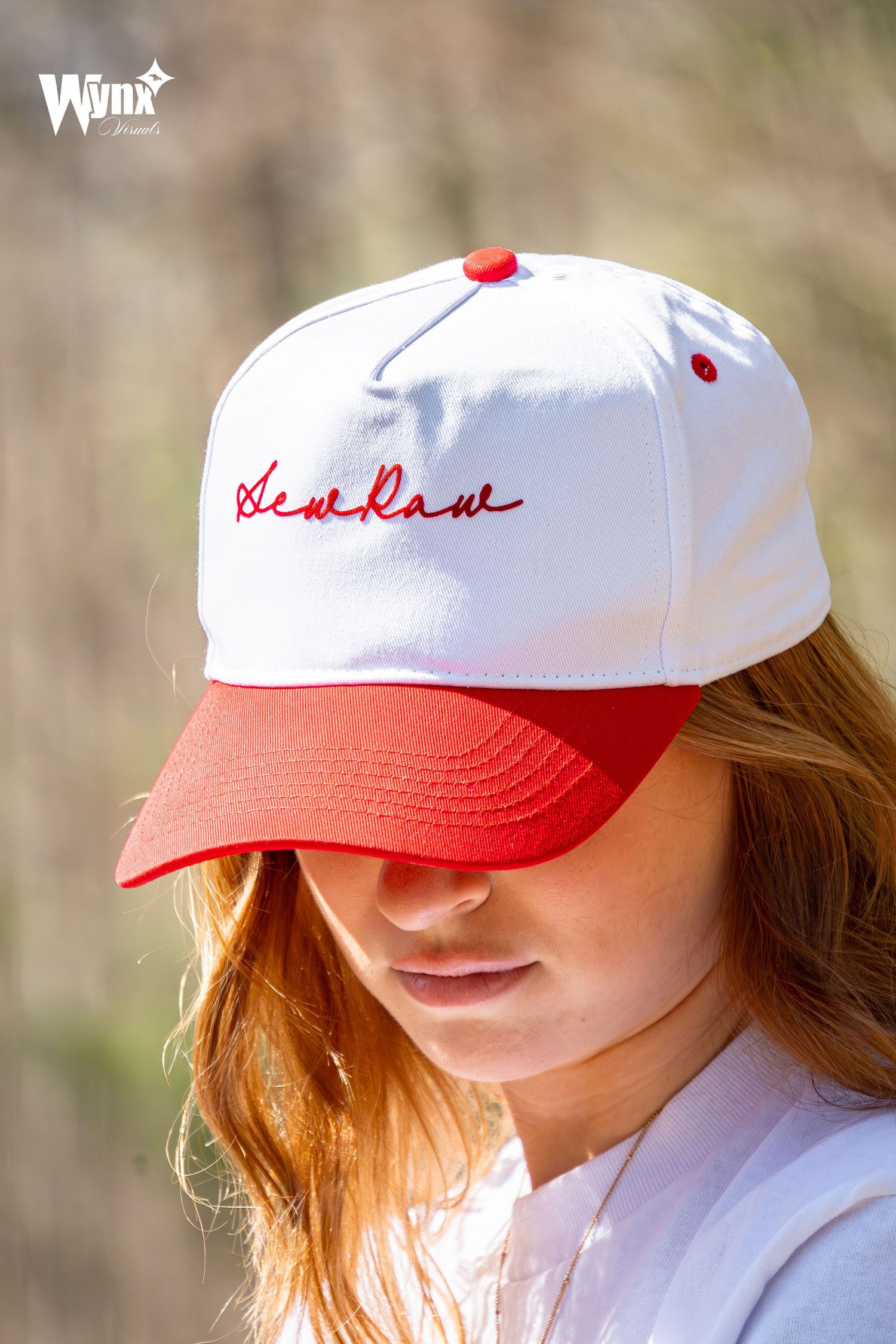 Close-up of a woman in a white and red Sewlaw script cap with a red brim, looking downward outdoors in soft sunlight.