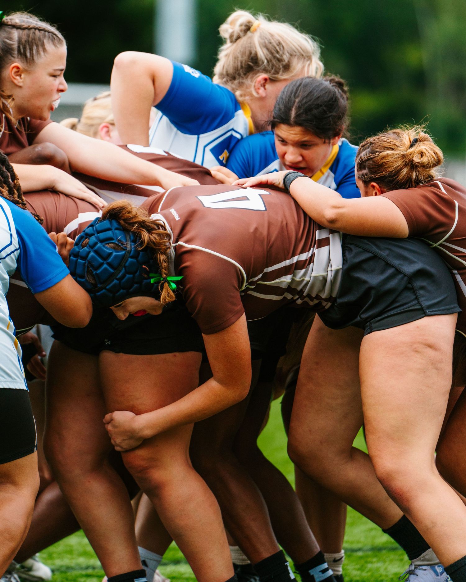 Women's rugby teams bind together in a tight scrum, with players in brown and blue jerseys pushing against each other.