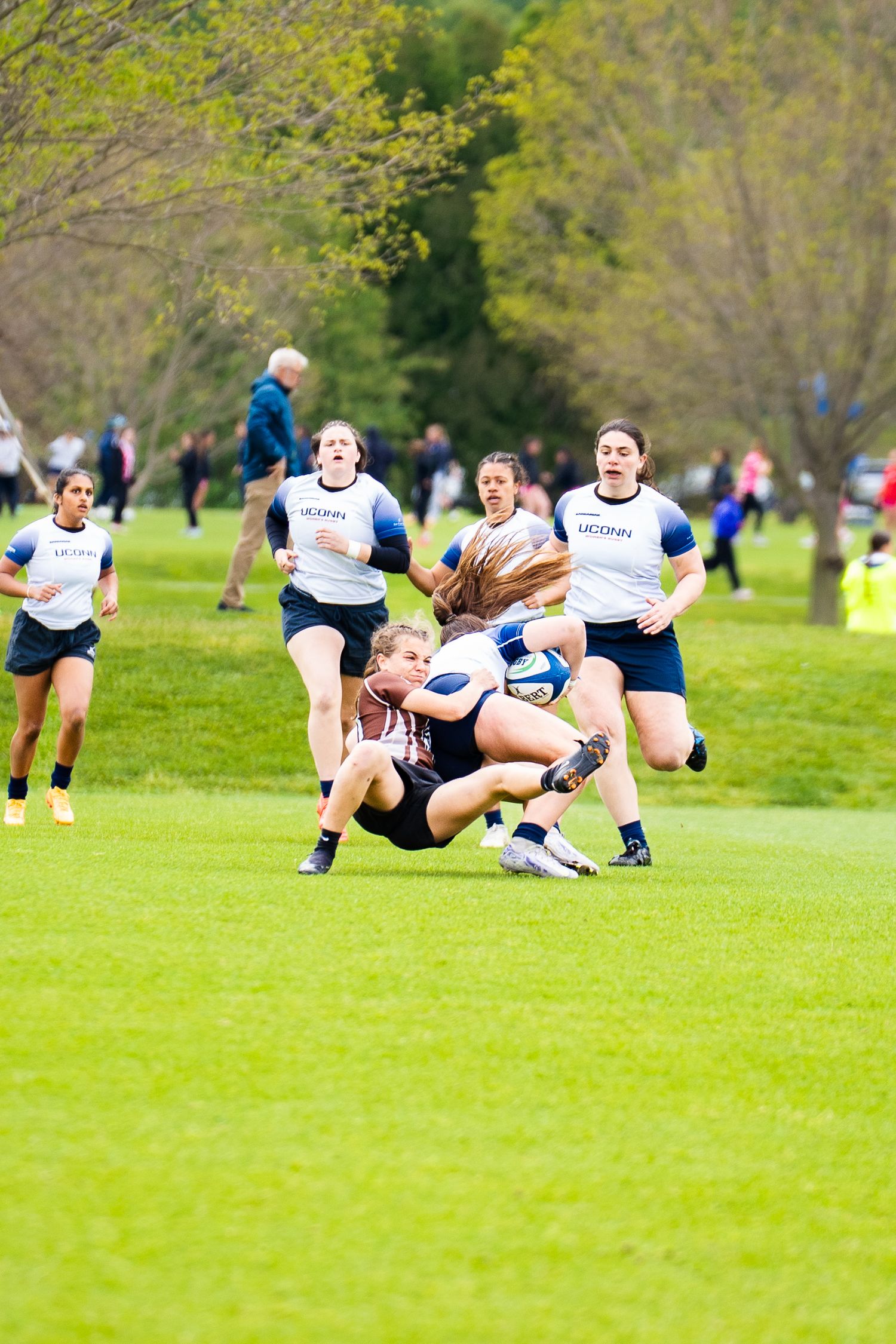 A UConn rugby player in white is tackled to the grass by a player in a brown jersey as teammates run in during a women's match.