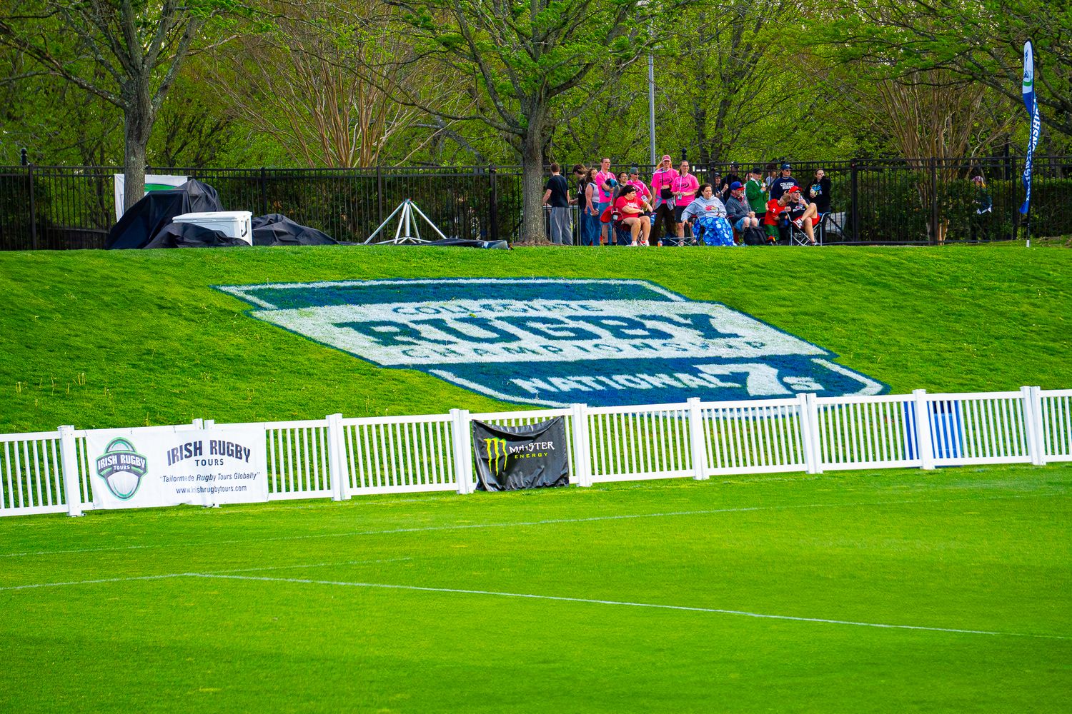 A rugby pitch with a logo on a grassy hillside, white fencing, and spectators gathered under trees on a spring day.