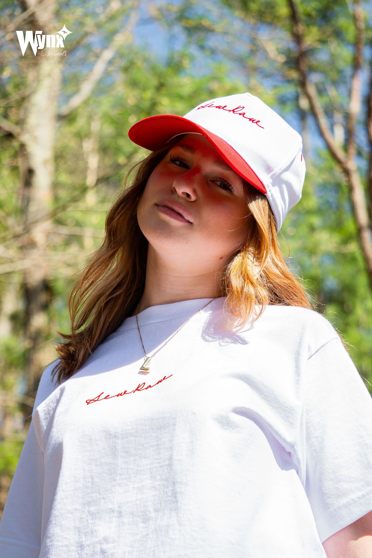 Woman tilting her head while wearing a white and red Sewlaw script cap and white tee with a pendant necklace outdoors.
