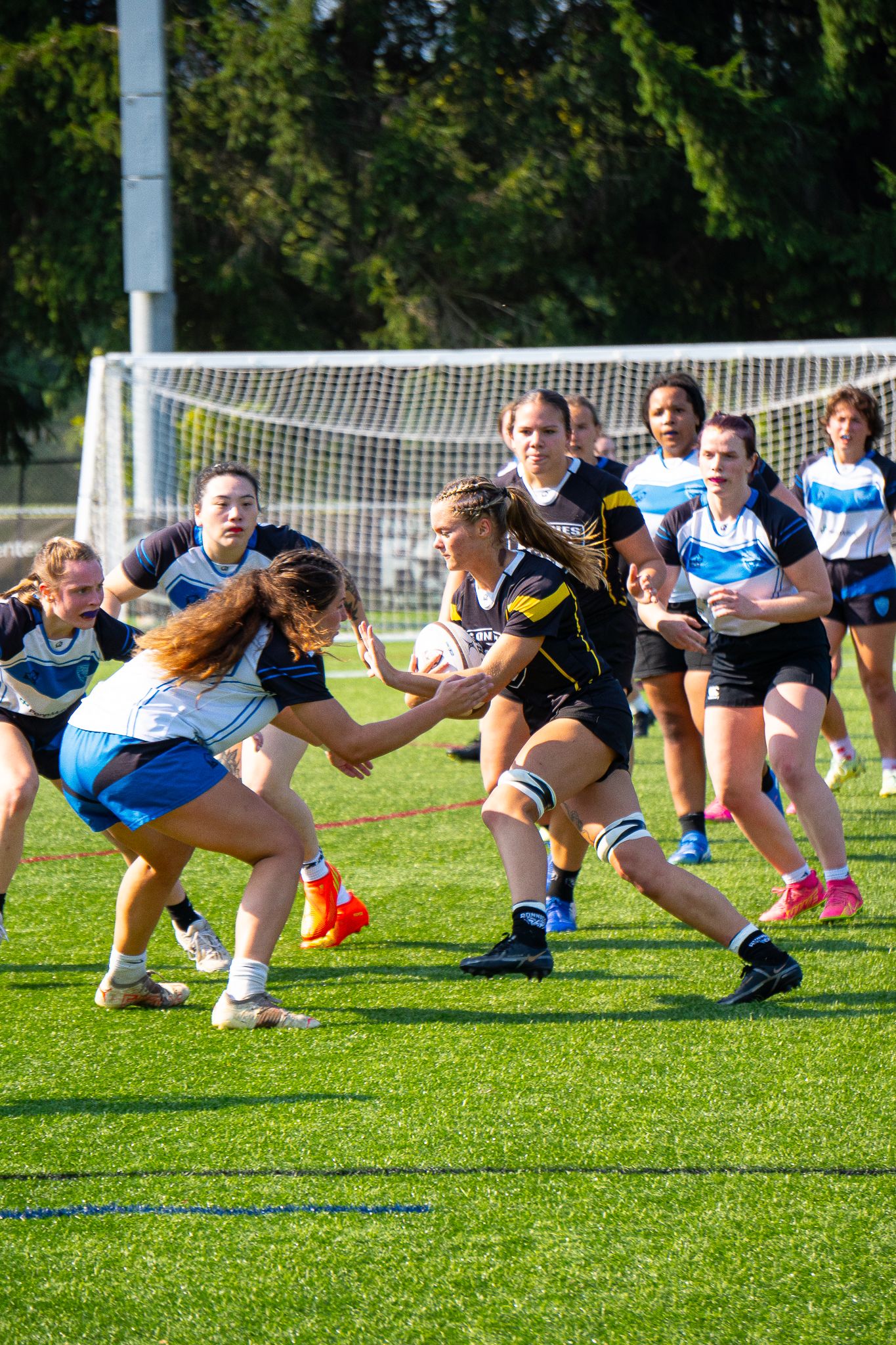 A women's rugby player in a black and gold uniform runs with the ball toward two defenders in blue and white on a turf field.