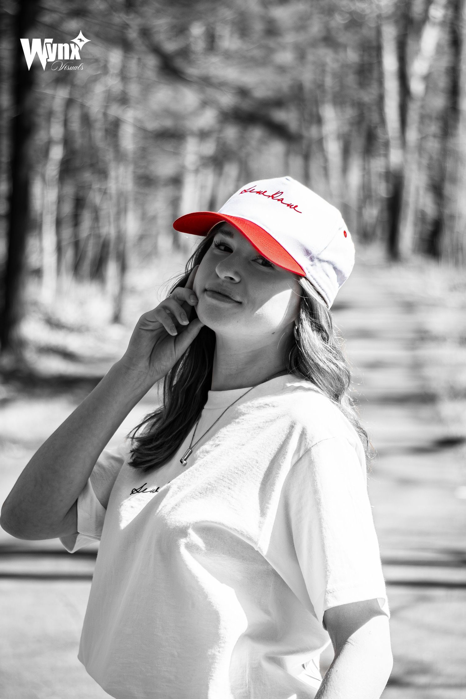 Black and white outdoor portrait of a woman touching her cheek, wearing a white tee and red-brim Sewlaw cap in selective red color.