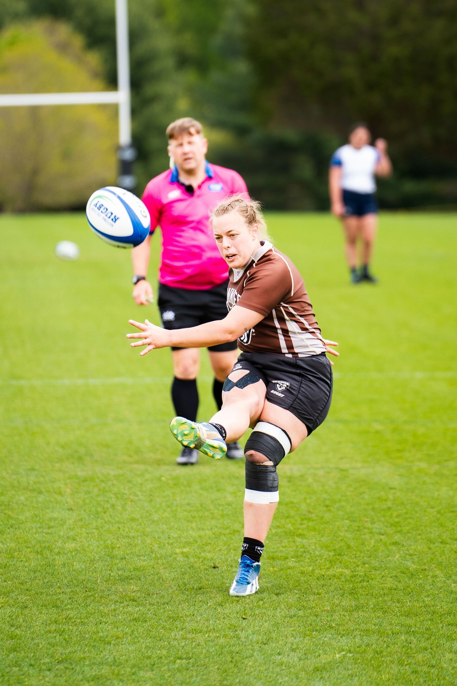 A women's rugby player in a brown jersey with a knee brace kicks the ball while a referee in pink watches on a green pitch.