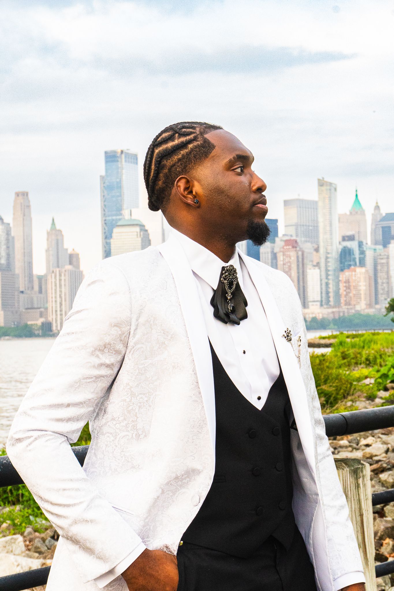 Man with braided hair in a white brocade tuxedo jacket, black vest and bow tie posing in profile with a city skyline behind him.