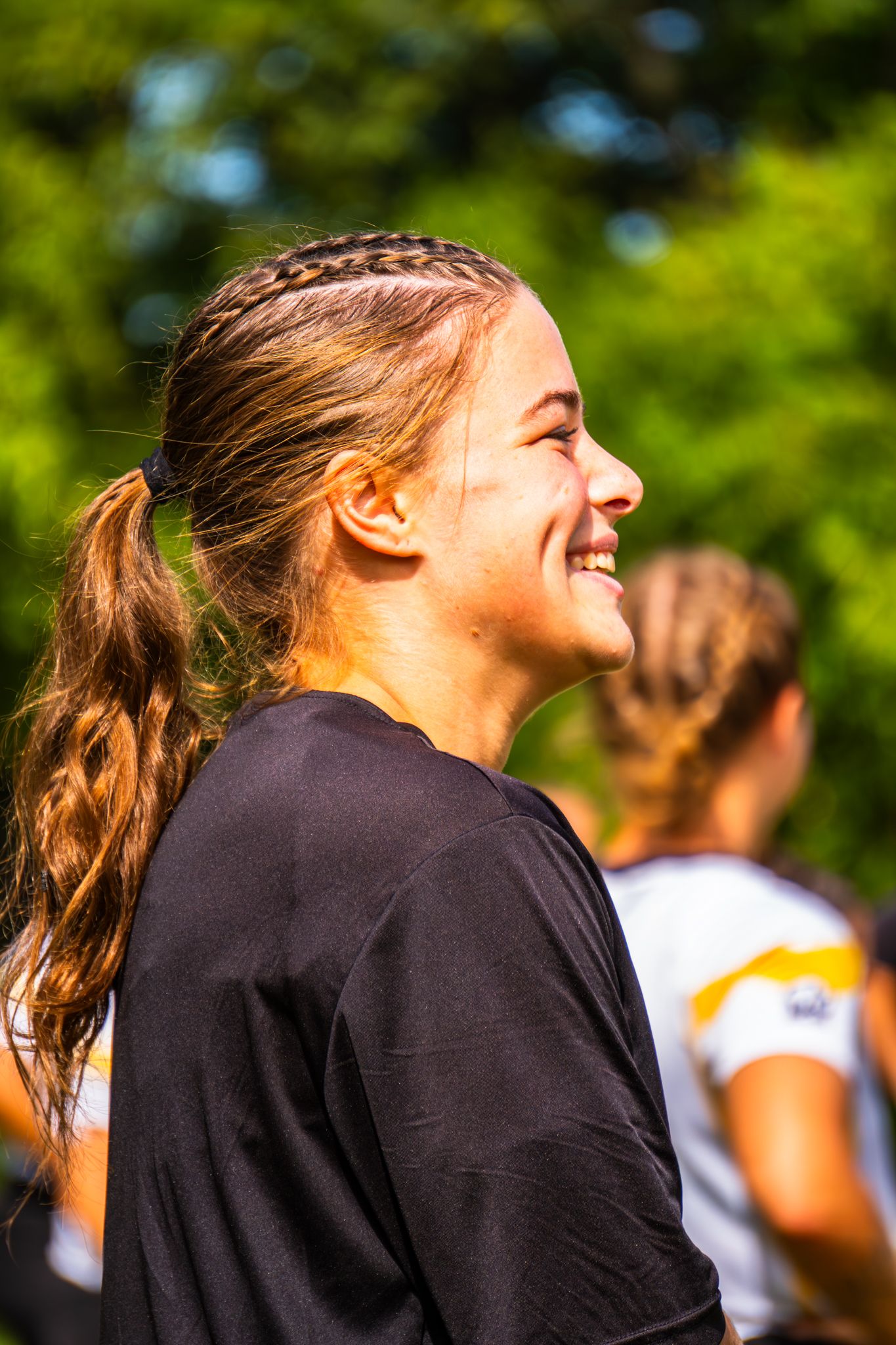 Smiling woman with a braided ponytail in a black shirt stands outdoors with green trees and a teammate blurred behind her.