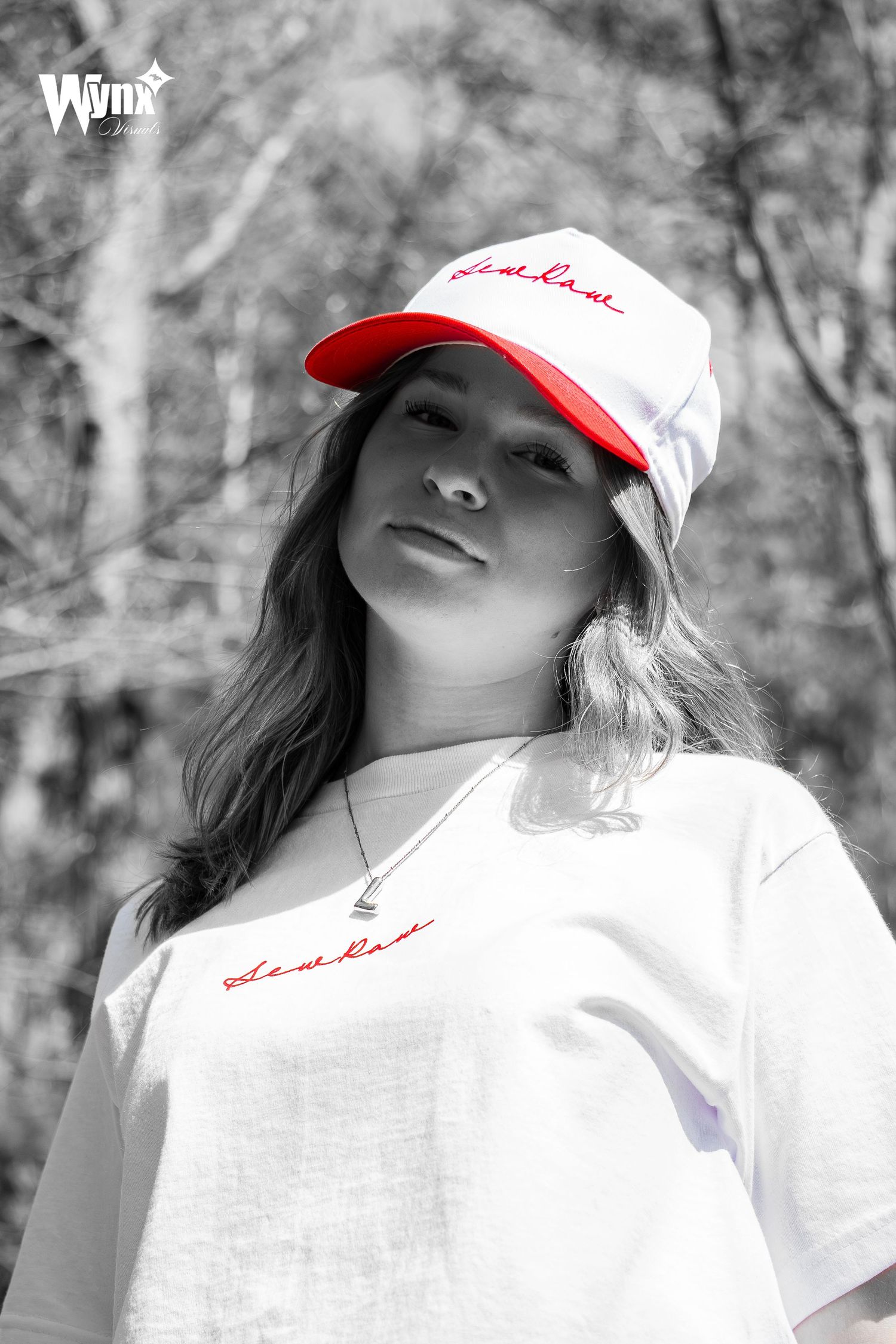 Black and white portrait of a woman in a white and red baseball cap and embroidered tee with red accents in the woods.