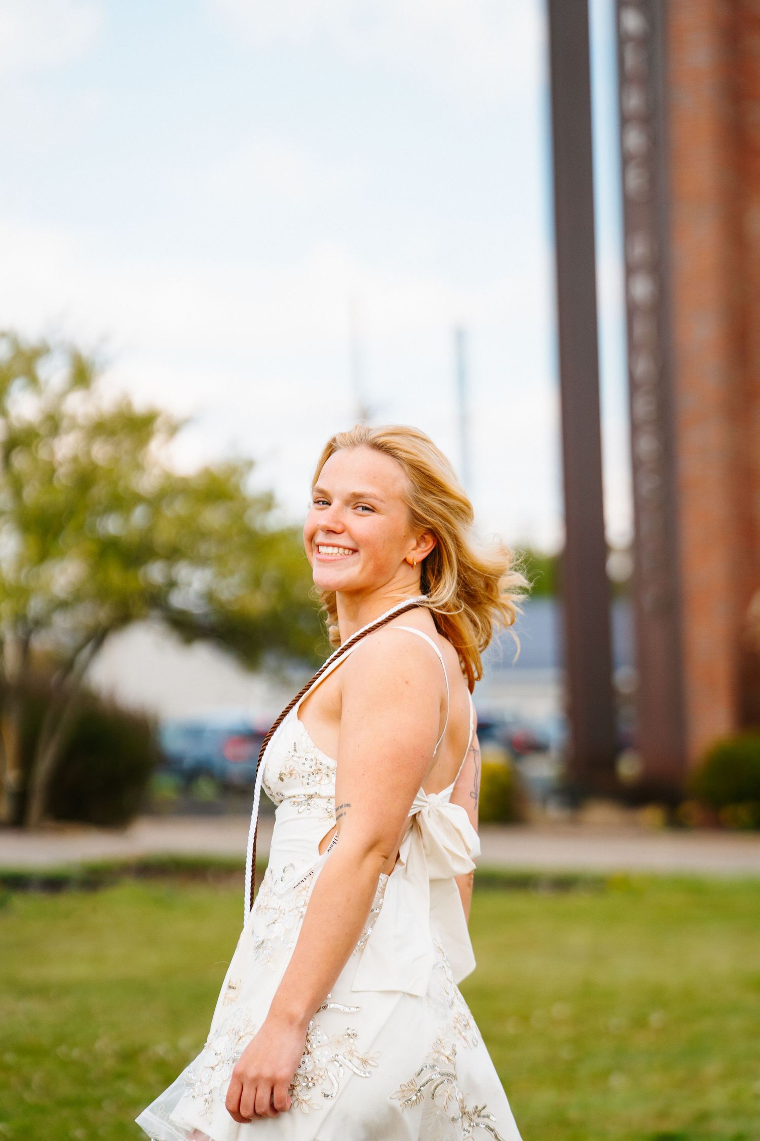 Smiling woman in a white lace dress looks over her shoulder on a grassy campus lawn in front of a tall brick tower.