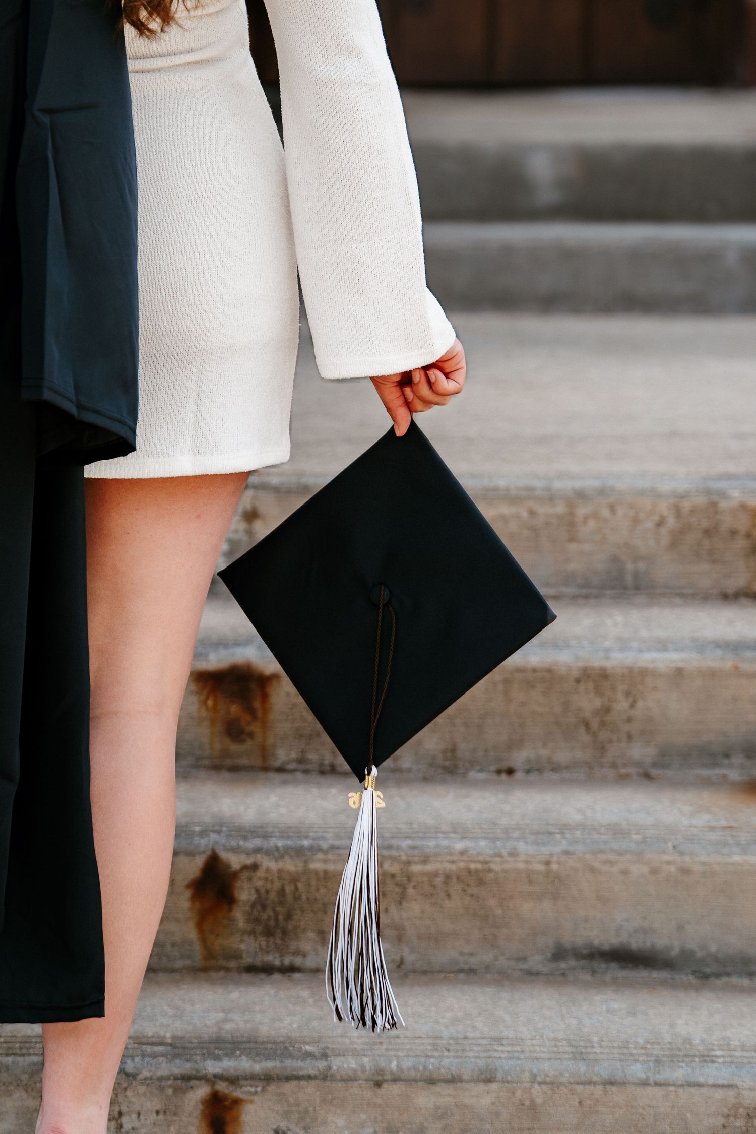 Graduate in a white dress holds a black mortarboard cap with a silver tassel while standing on weathered stone steps.