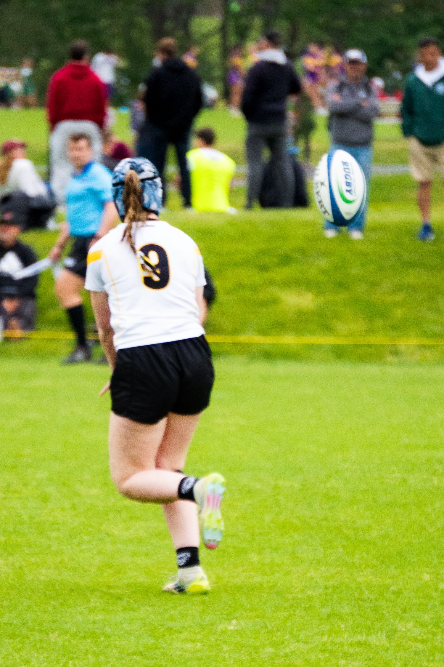 A rugby player in a white jersey and blue scrum cap runs downfield as a rugby ball and spectators appear in the background.