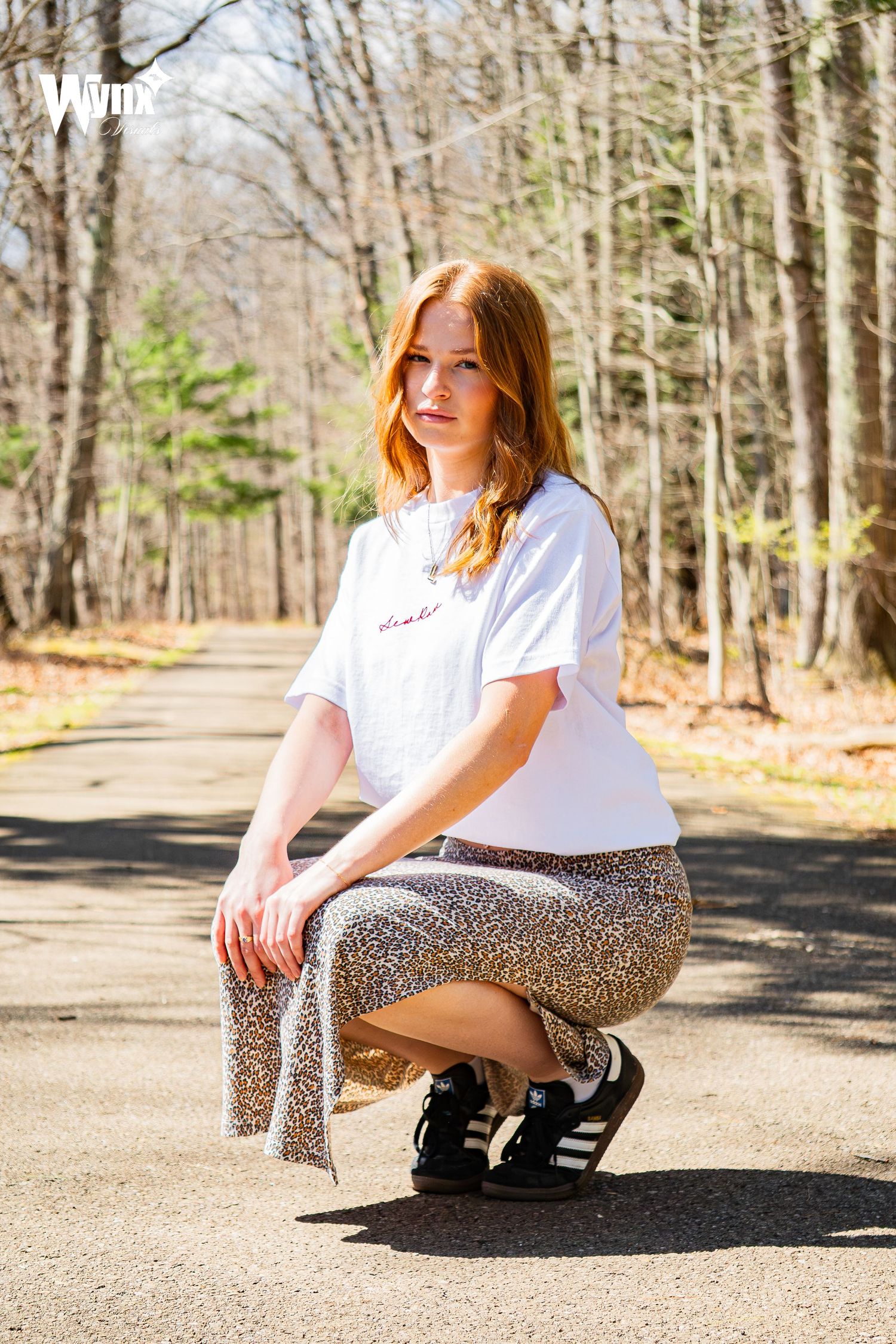 Woman crouches on a wooded path in a white Sewlaw tee, leopard print skirt and black Adidas sneakers on a sunny spring day.