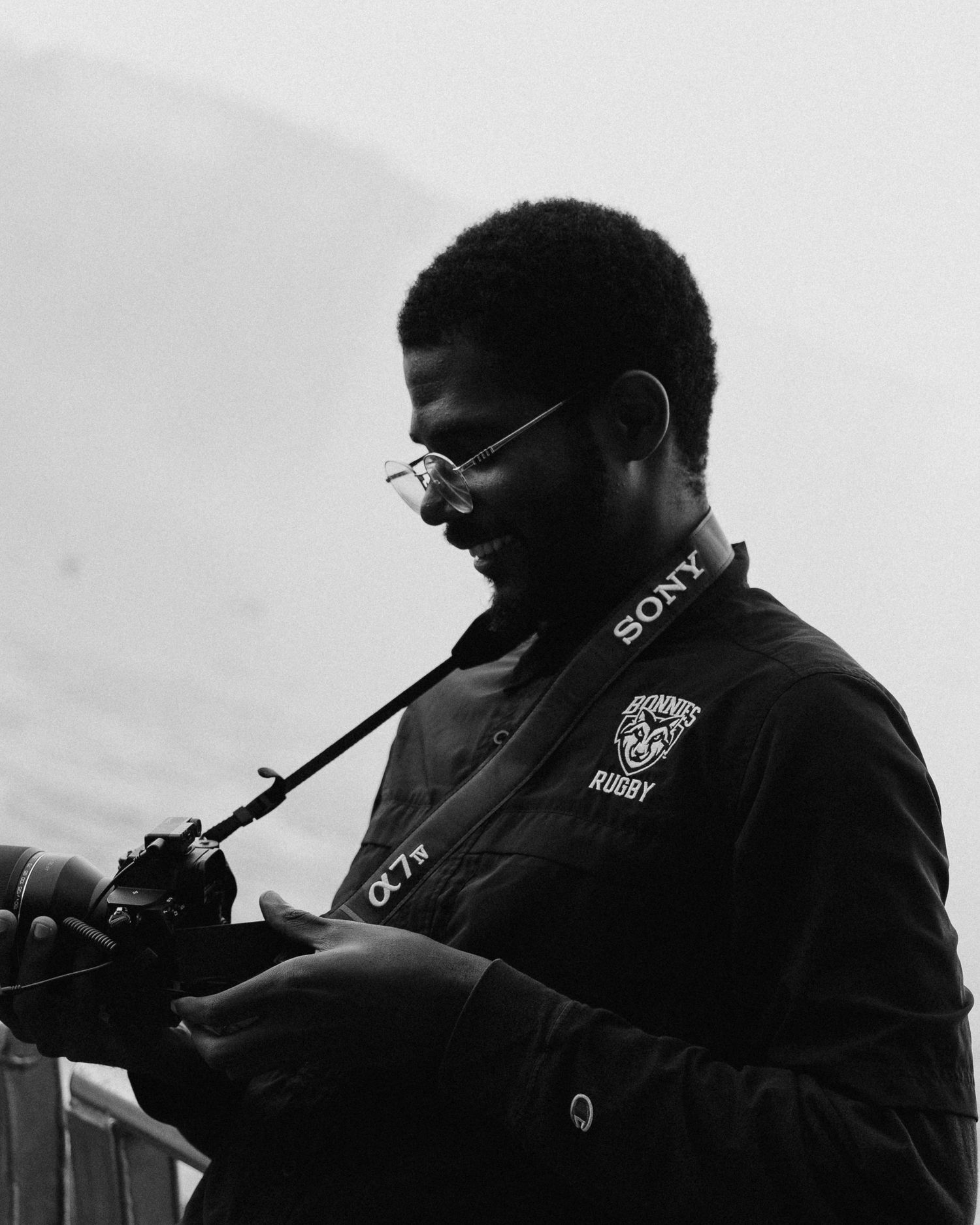 Black and white portrait of a smiling man with glasses looking down at a Sony camera, wearing a rugby jacket in a misty outdoor setting.