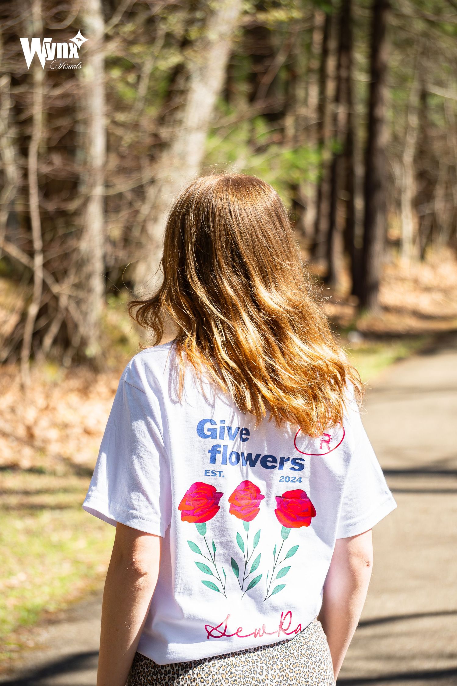 Back view of a woman on a wooded trail wearing a white Give Flowers tee with red poppy graphics and a leopard print skirt.