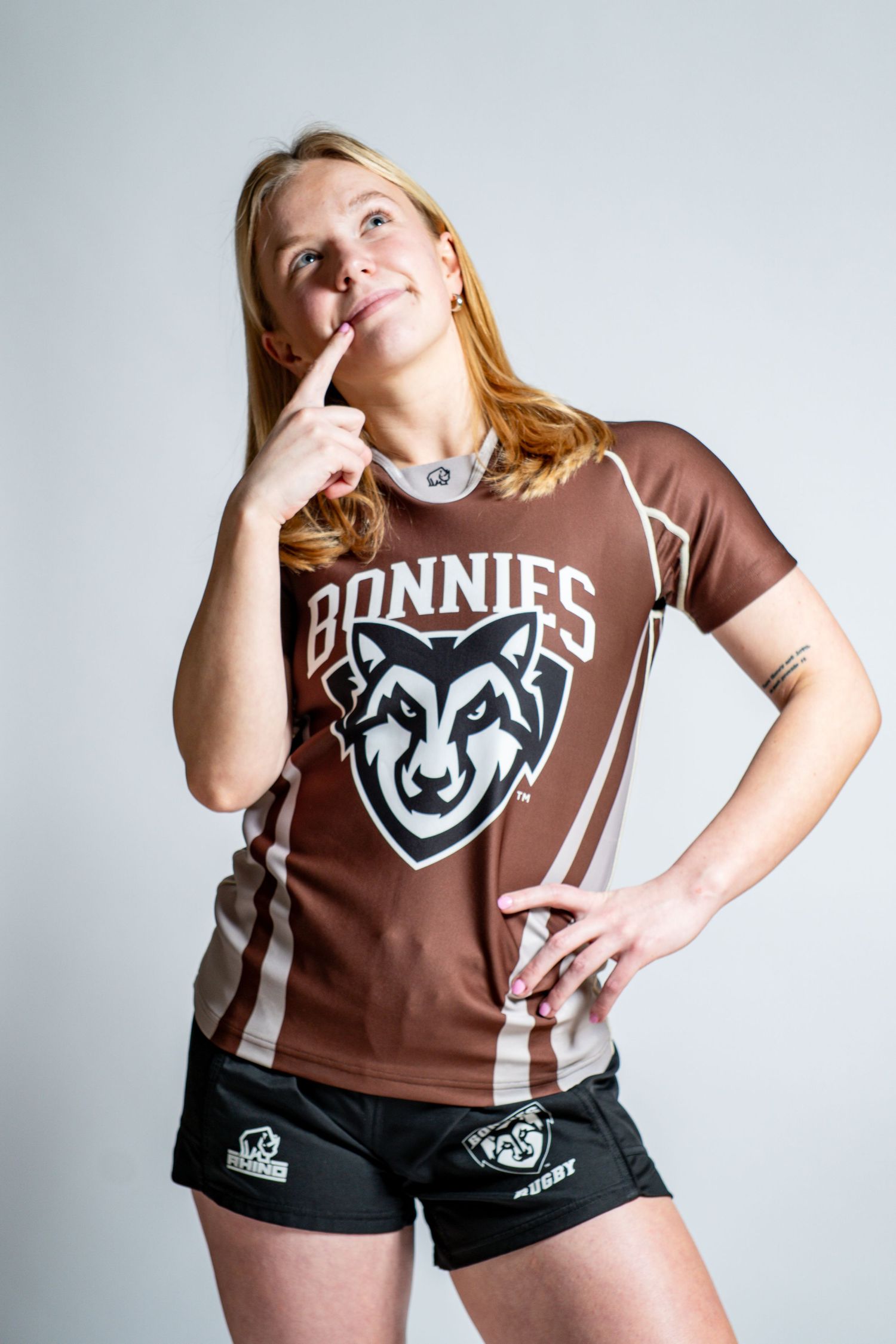 Rugby player in a brown Bonnies wolf logo jersey and black shorts strikes a thoughtful pose with a finger to her chin.
