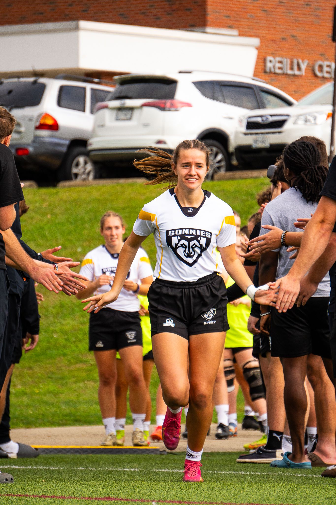 A smiling women's rugby player in a white Bonnies jersey jogs onto the field as teammates and staff reach out to greet her.