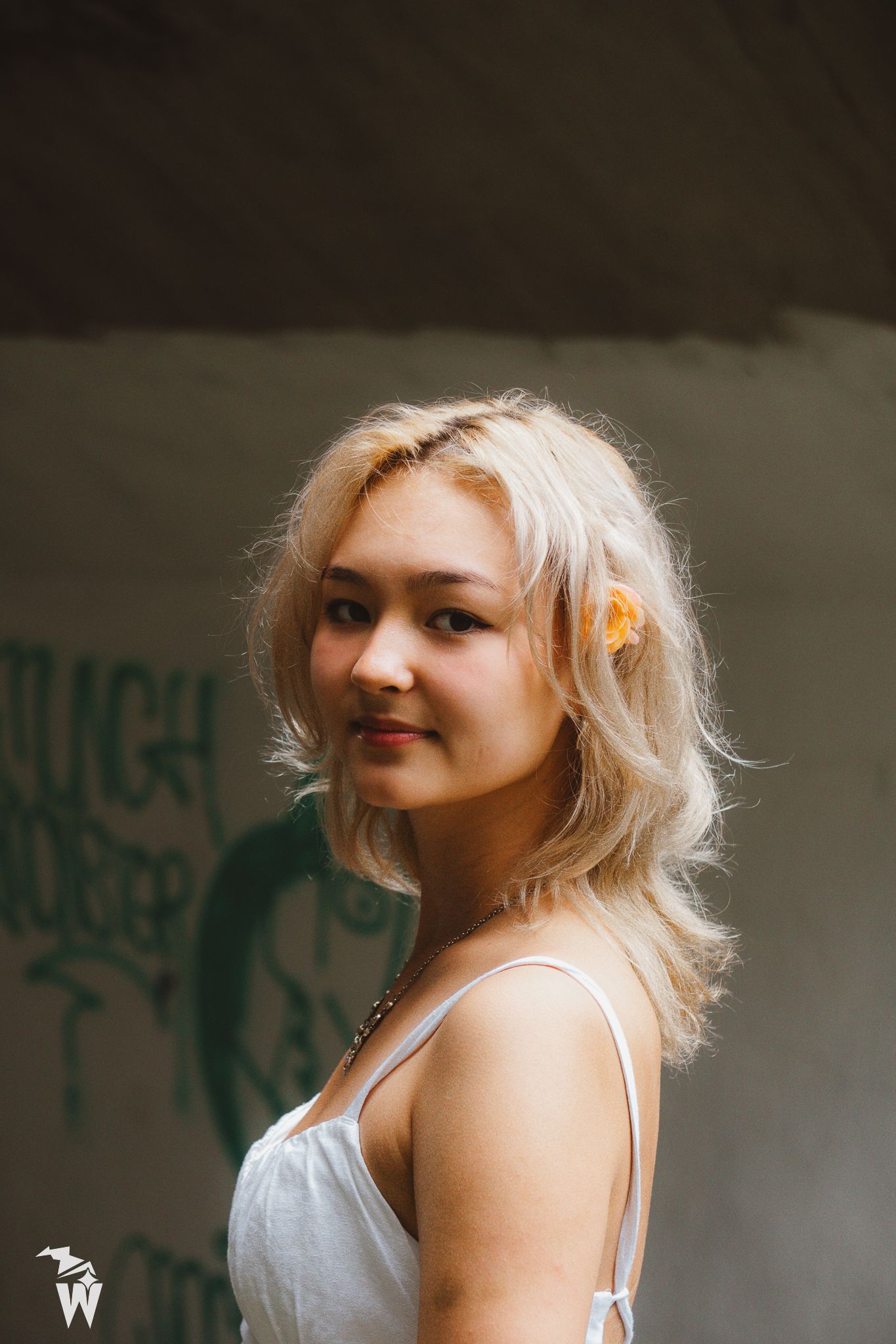 Blonde woman in a white dress with an orange flower in her hair looks over her shoulder in front of a graffiti wall.