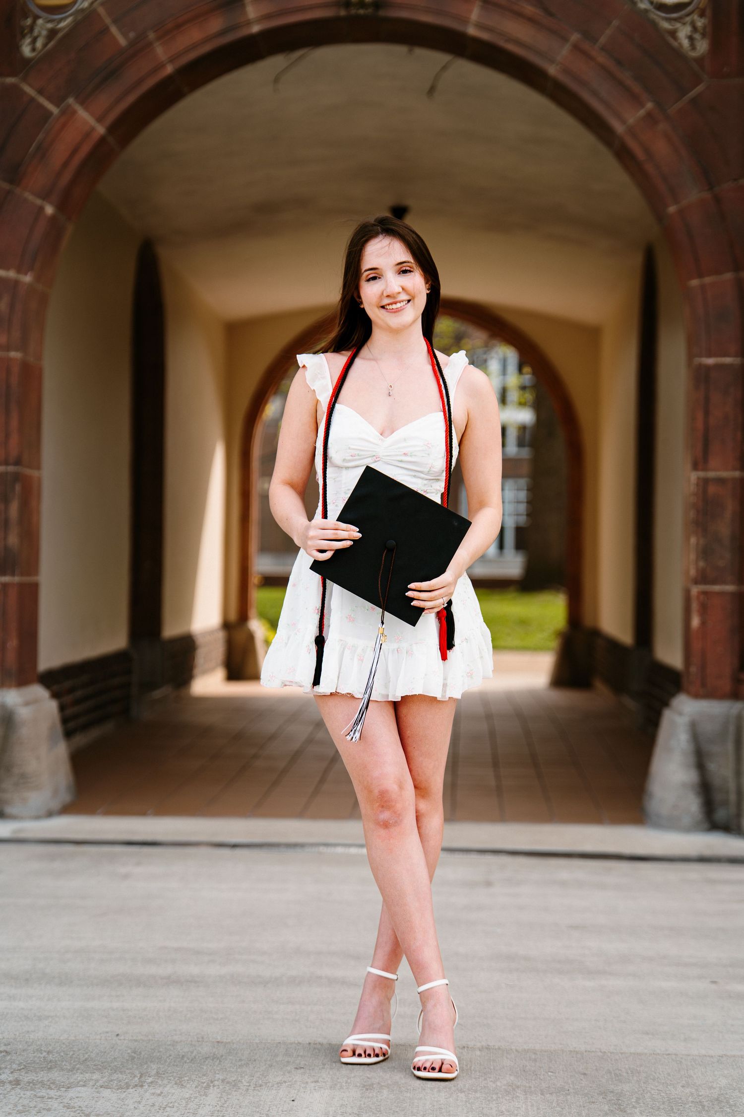 A smiling graduate in a white dress and red-trimmed stole holds her black cap while standing beneath a brick archway on campus.