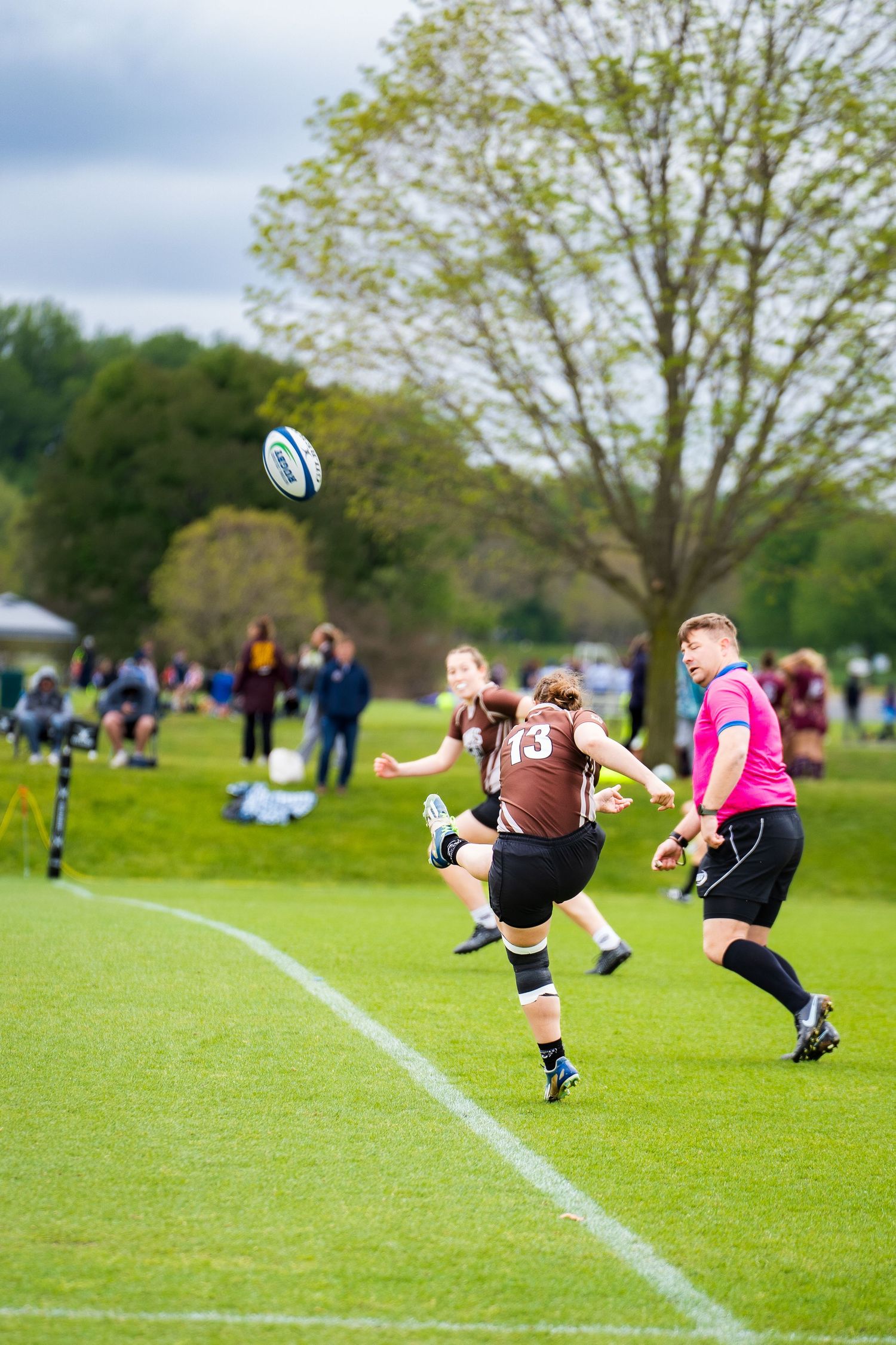 A rugby player in a brown jersey kicks the ball high into the air as a referee in pink and spectators watch from the sideline.